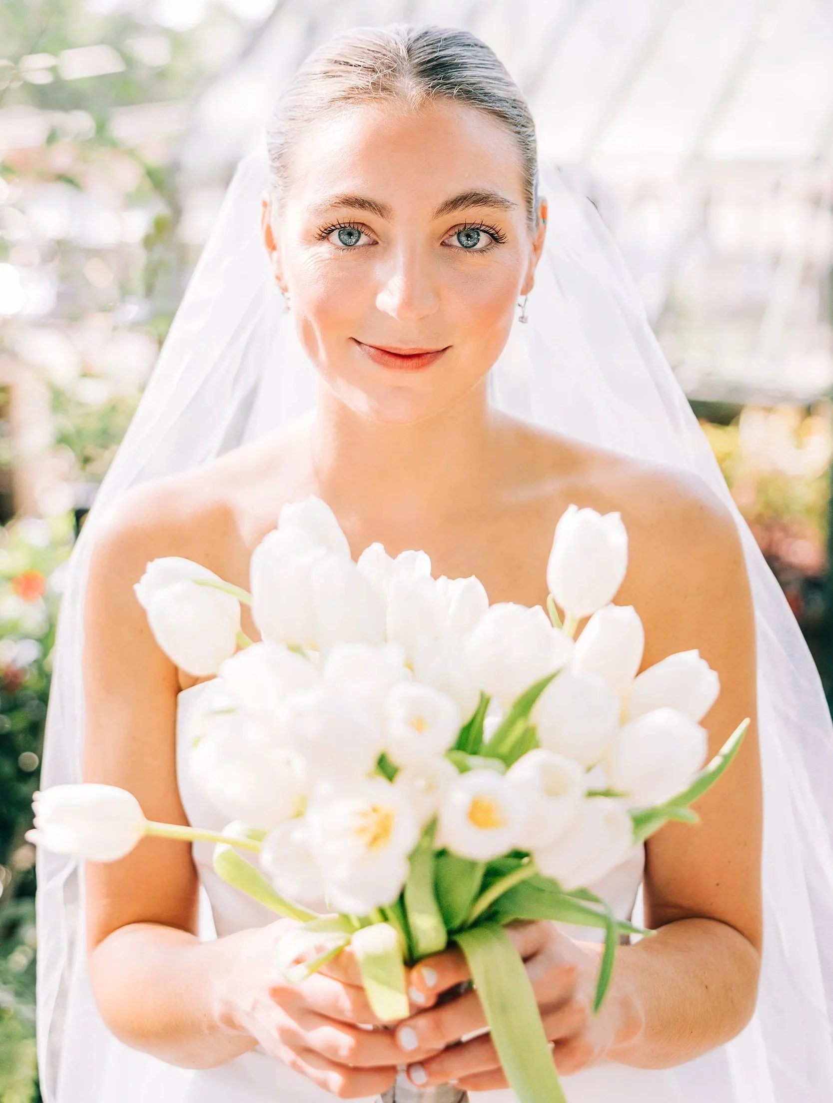 A bride in a wedding dress holding a bouquet of white flowers outdoors with a bright, blurred background.