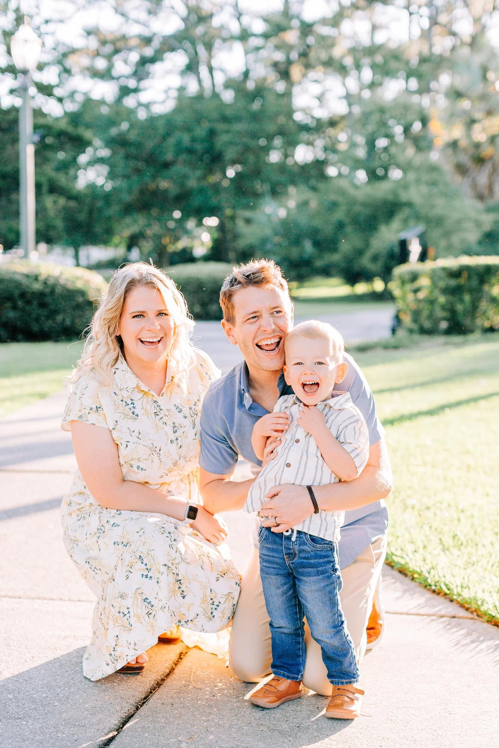 A family of four, a woman, a man, and two young boys, smiling and posing outdoors on a sunny day in a park or neighborhood with green trees and bushes in the background.
