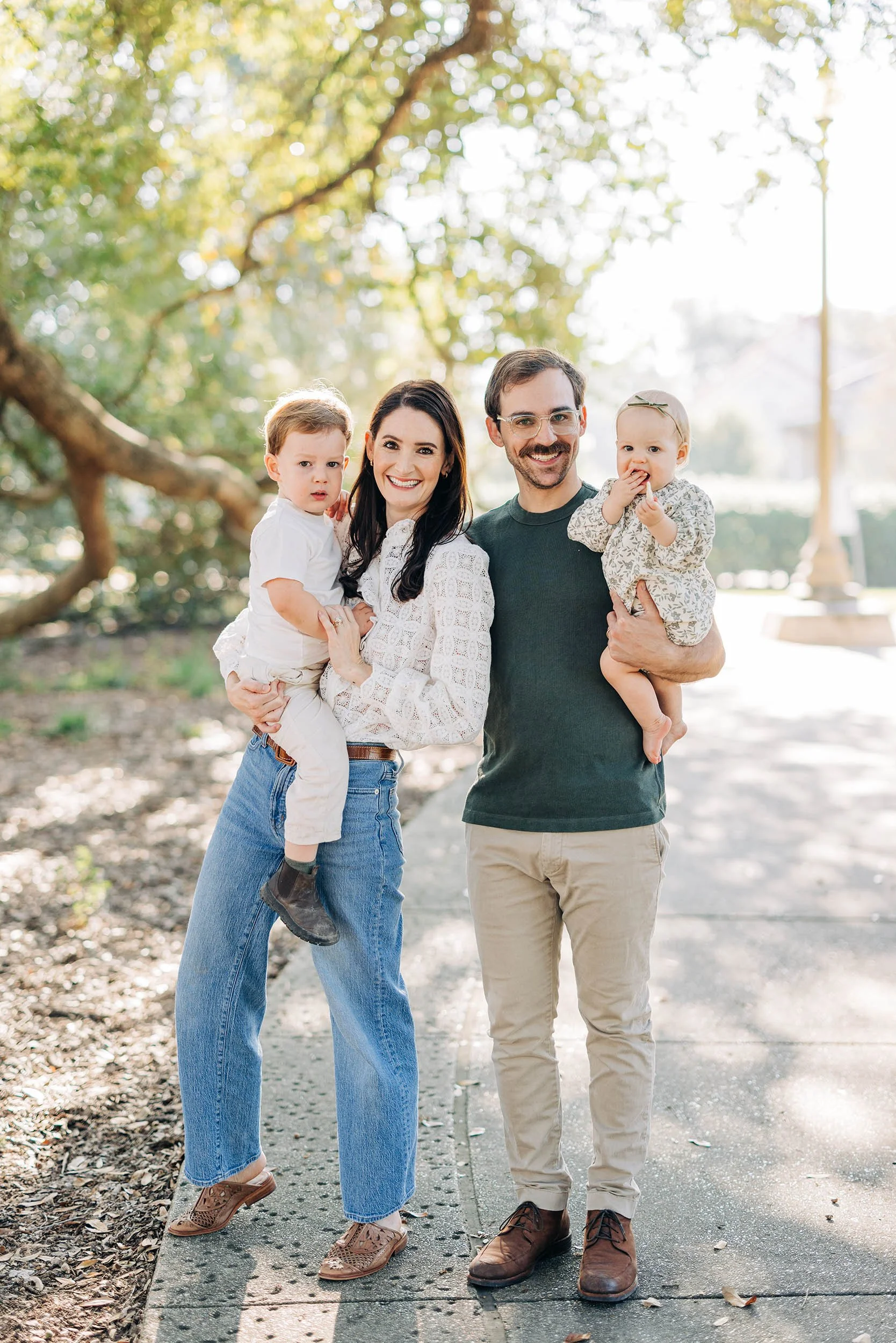 A happy family of four standing outdoors on a sidewalk, with the mother holding a young boy, and the father holding a baby girl, both children appearing to be around 2 years old. The background includes trees and sunlight.