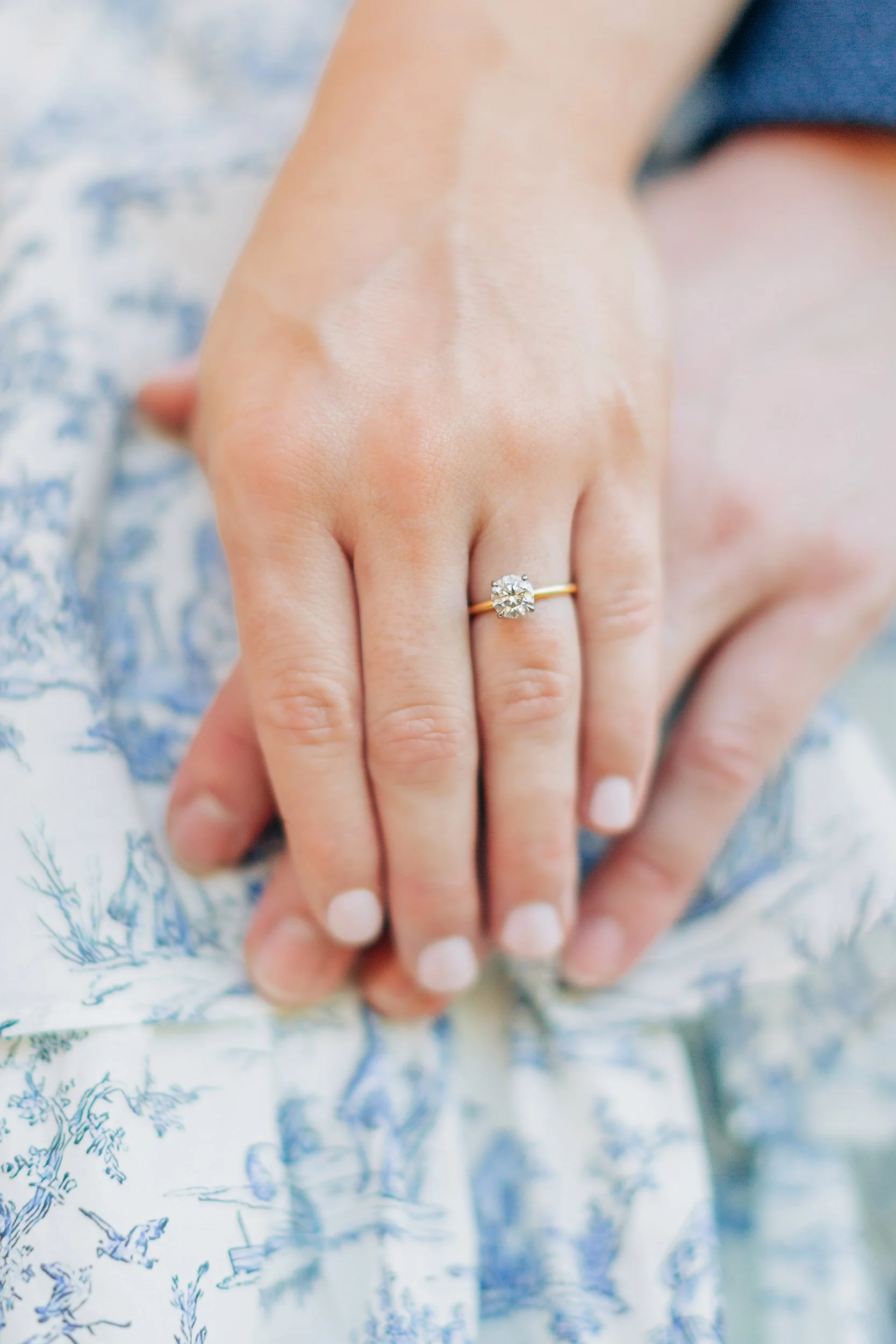 Close-up of a person showing their left hand with an engagement ring featuring a large round cut diamond on a thin yellow gold band, resting on patterned fabric.