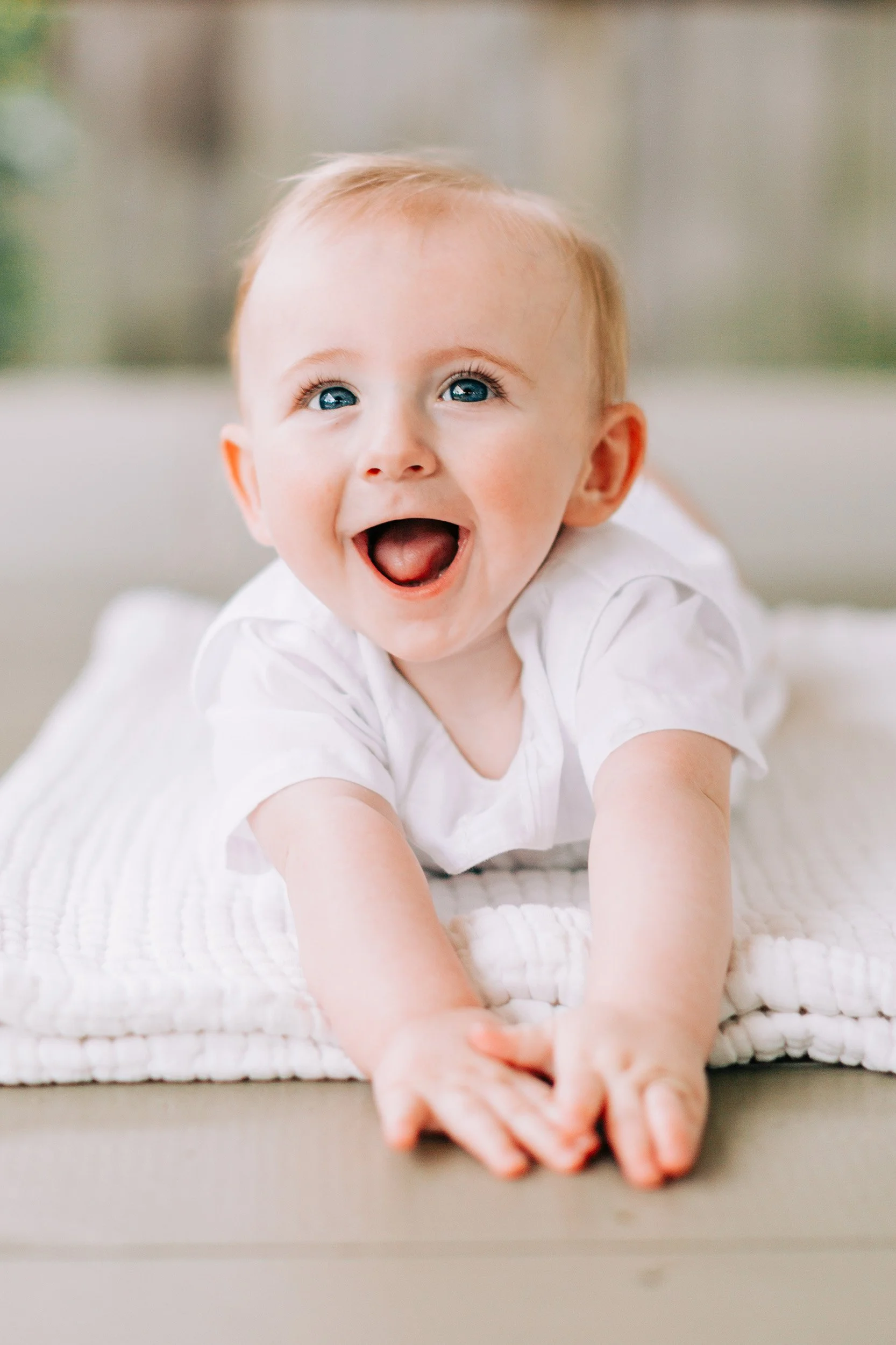 A happy baby with blue eyes and light hair, lying on a white blanket, with arms extended forward and mouth open in a smile.