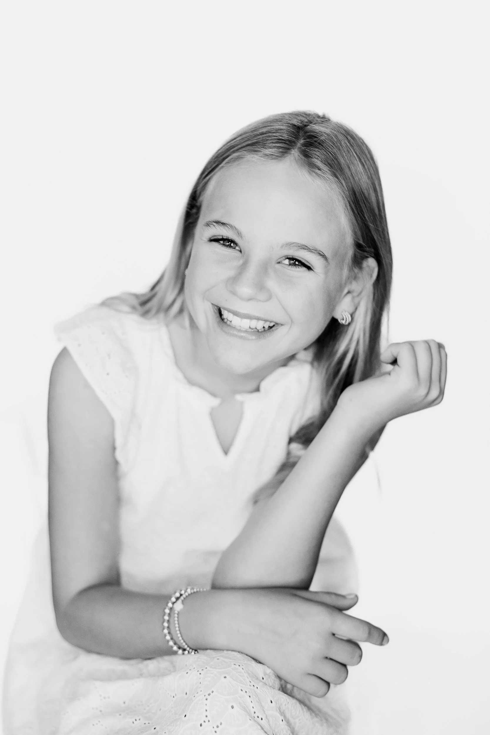 Black and white photo of a young girl smiling, resting her head on her hand, wearing a sleeveless dress and earrings, with a bracelet on her wrist.