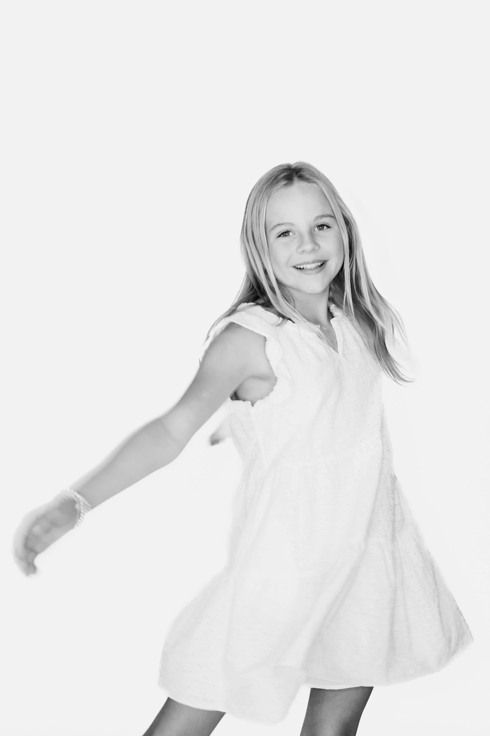 A young girl smiling and posing with her arms stretched out, wearing a white dress and accessories, against a plain light background.