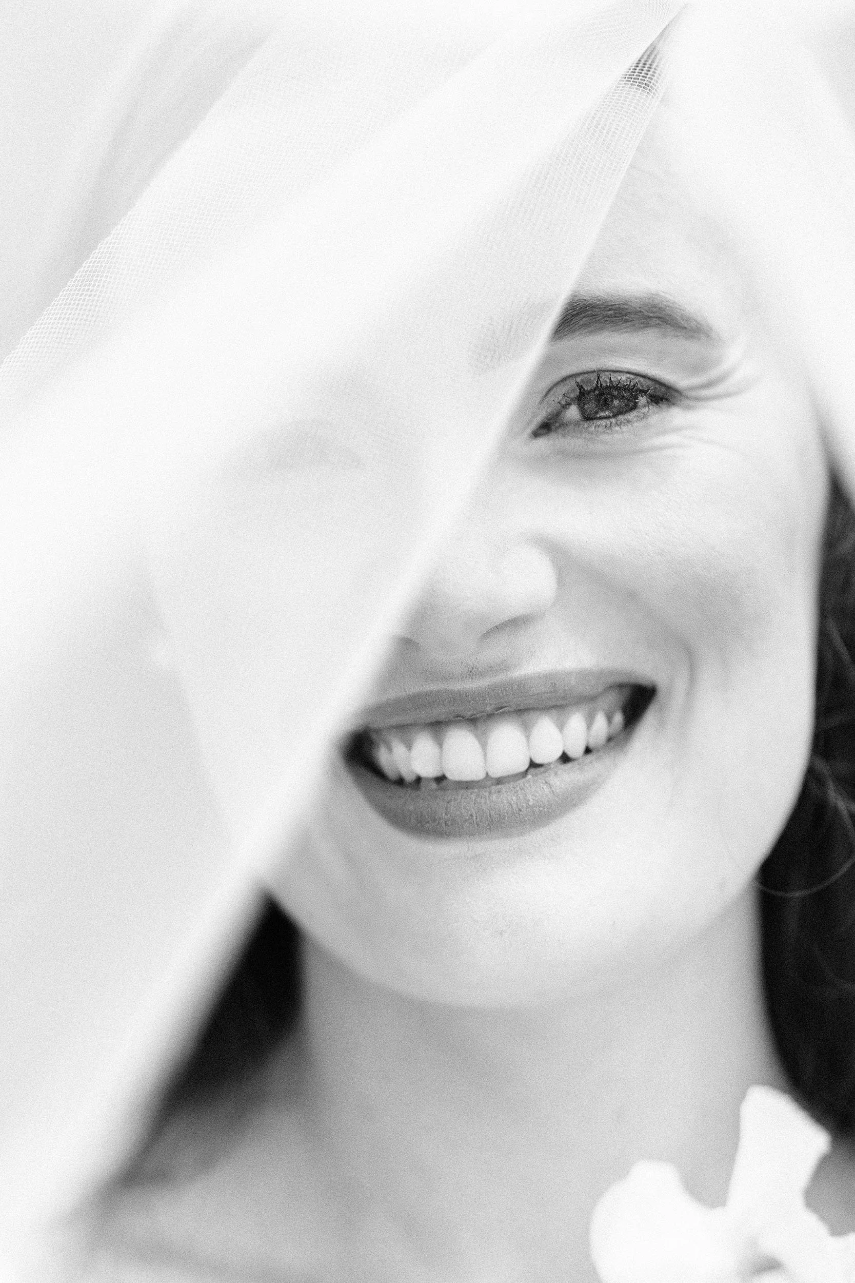 Close-up black and white photograph of a smiling woman with partial face covered by a veil, showing her eye and teeth.