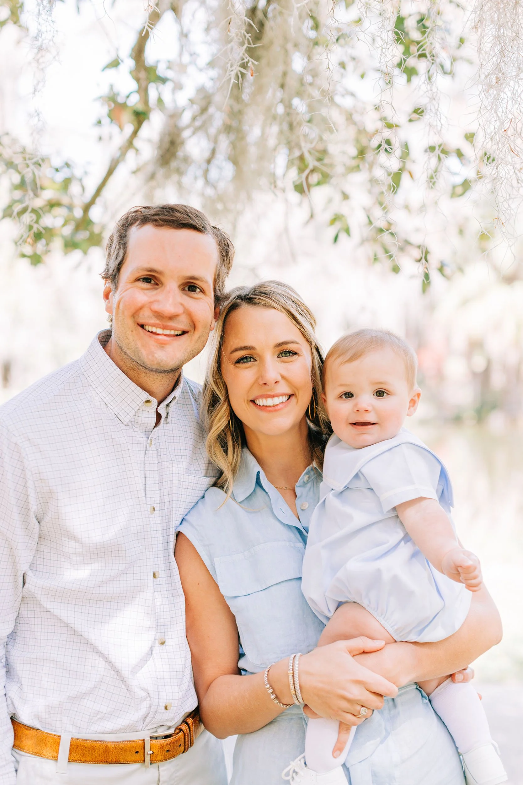 A smiling family of three standing outdoors with sunlight filtering through trees in the background. A man, woman, and young child are close together, with the woman holding the child.
