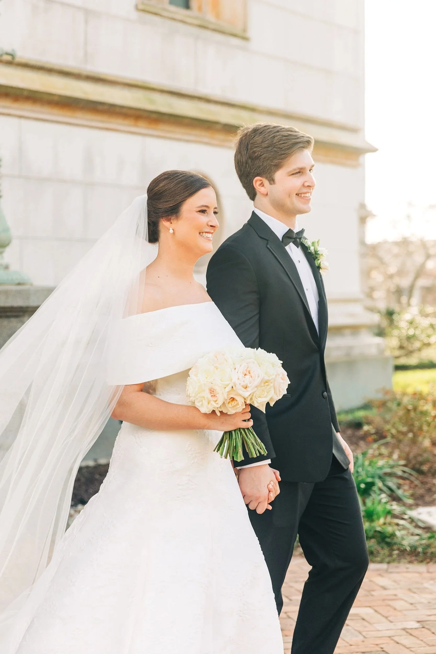 A bride and groom holding hands outdoors, smiling at each other, dressed in wedding attire, with the bride wearing a white gown and veil and the groom in a black tuxedo, against a background of a stone building and trees.