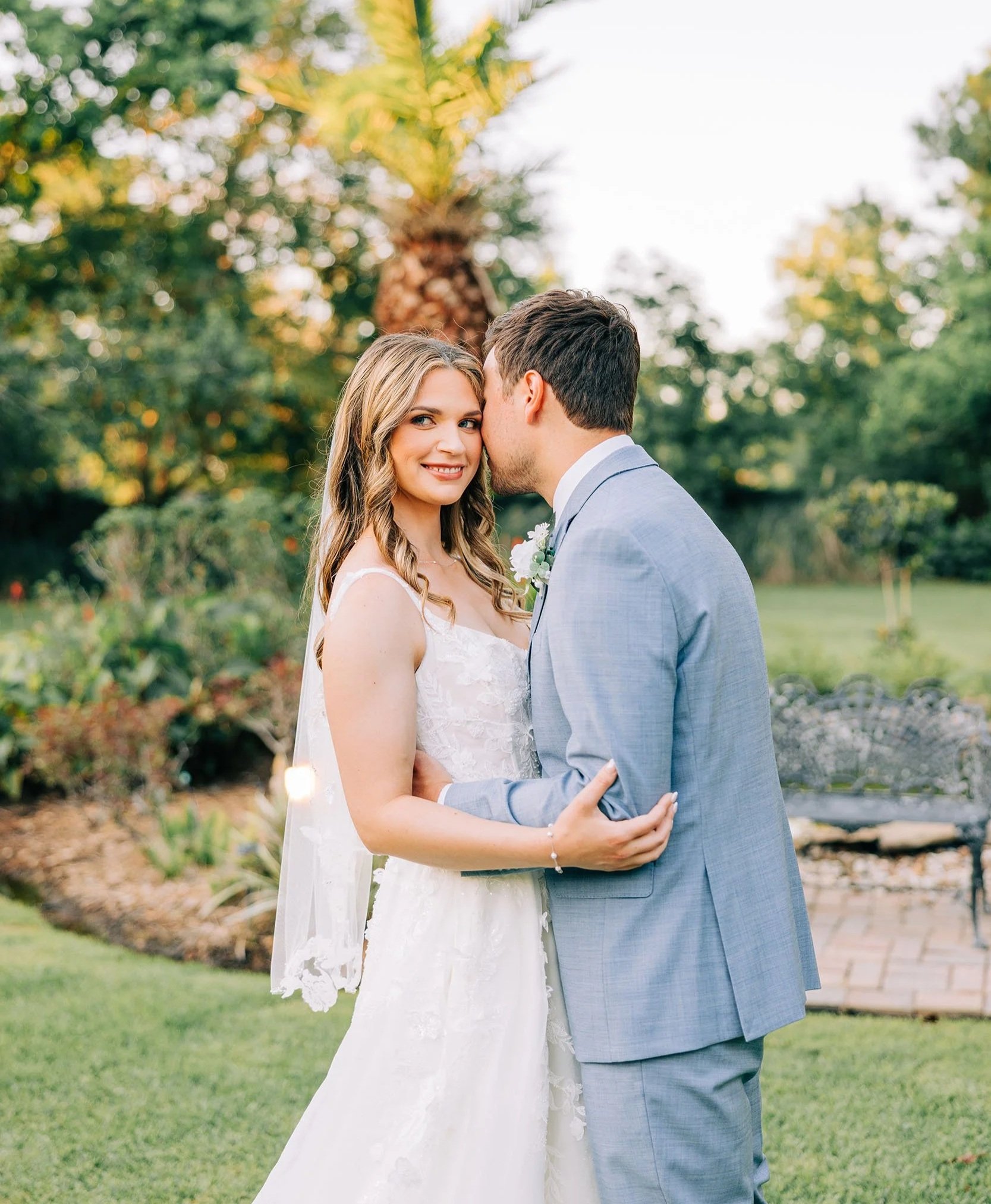 A bride and groom are embracing outdoors during their wedding, with a tropical tree and greenery in the background. The woman is wearing a white wedding dress with lace details and has long, wavy blonde hair. The man is dressed in a light gray suit with a white shirt and has dark hair. The groom is whispering or kissing the bride's temple, and she is smiling softly.