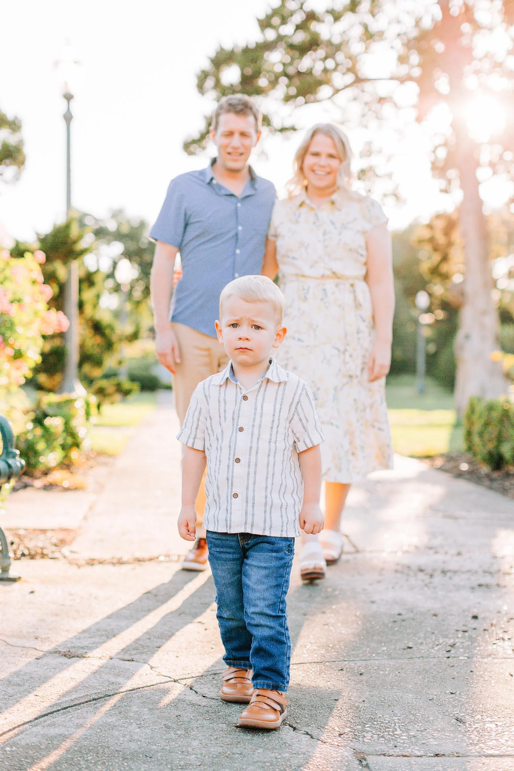 A young boy walking on a paved path in a park during sunset, with a happy family in the background.