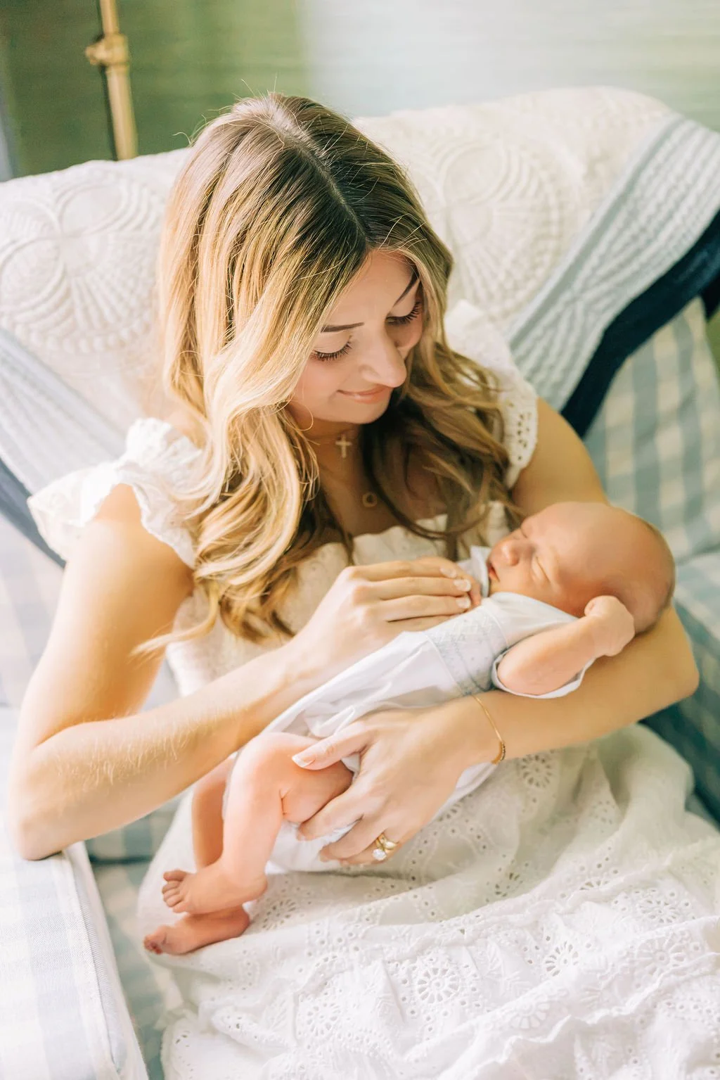A woman cradles a newborn baby, looking at the baby lovingly. The woman is sitting on a bed, wearing a white dress with lace details, and the baby is dressed in a white outfit.