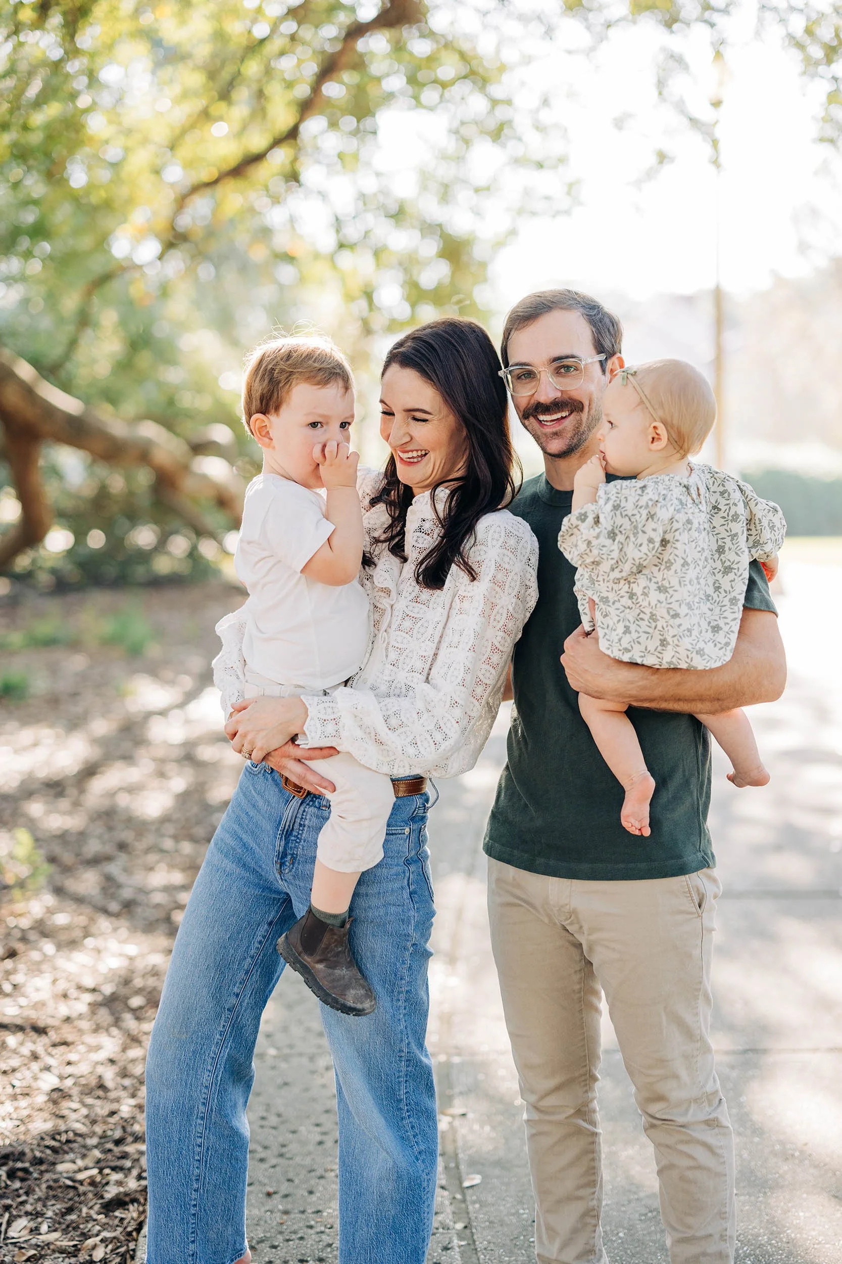 A family of four standing outdoors in a park-like setting with trees and sunlight in the background, smiling and enjoying their time together.