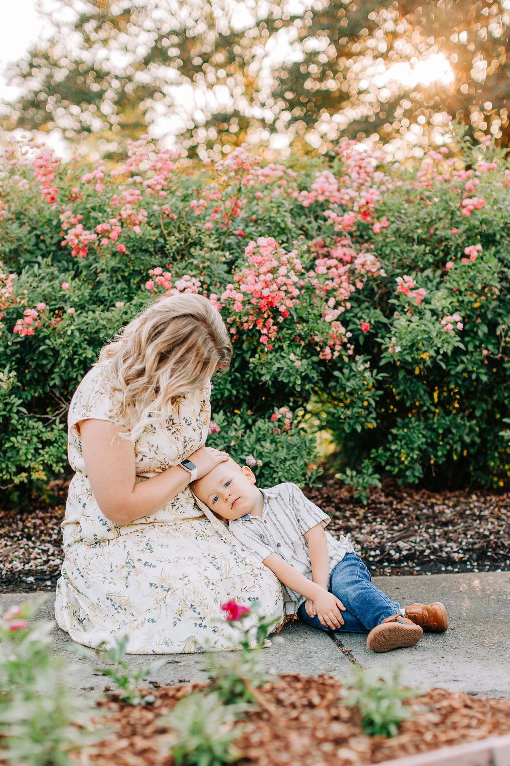 A woman with blonde hair sitting outdoors with a young boy. The woman is gently holding the boy's head, who is leaning on her lap. They are surrounded by pink flowering bushes and appear to be in a park or garden setting during sunset.