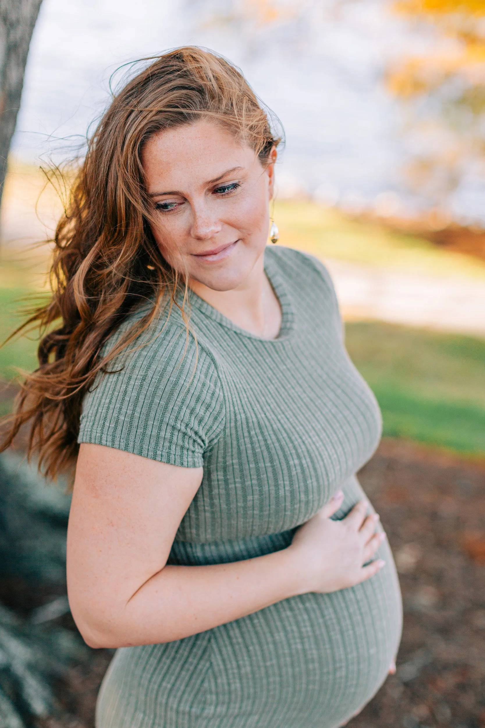 A pregnant woman with red hair and freckles outdoors, wearing a green ribbed dress, holding her belly, with a blurred autumn background.