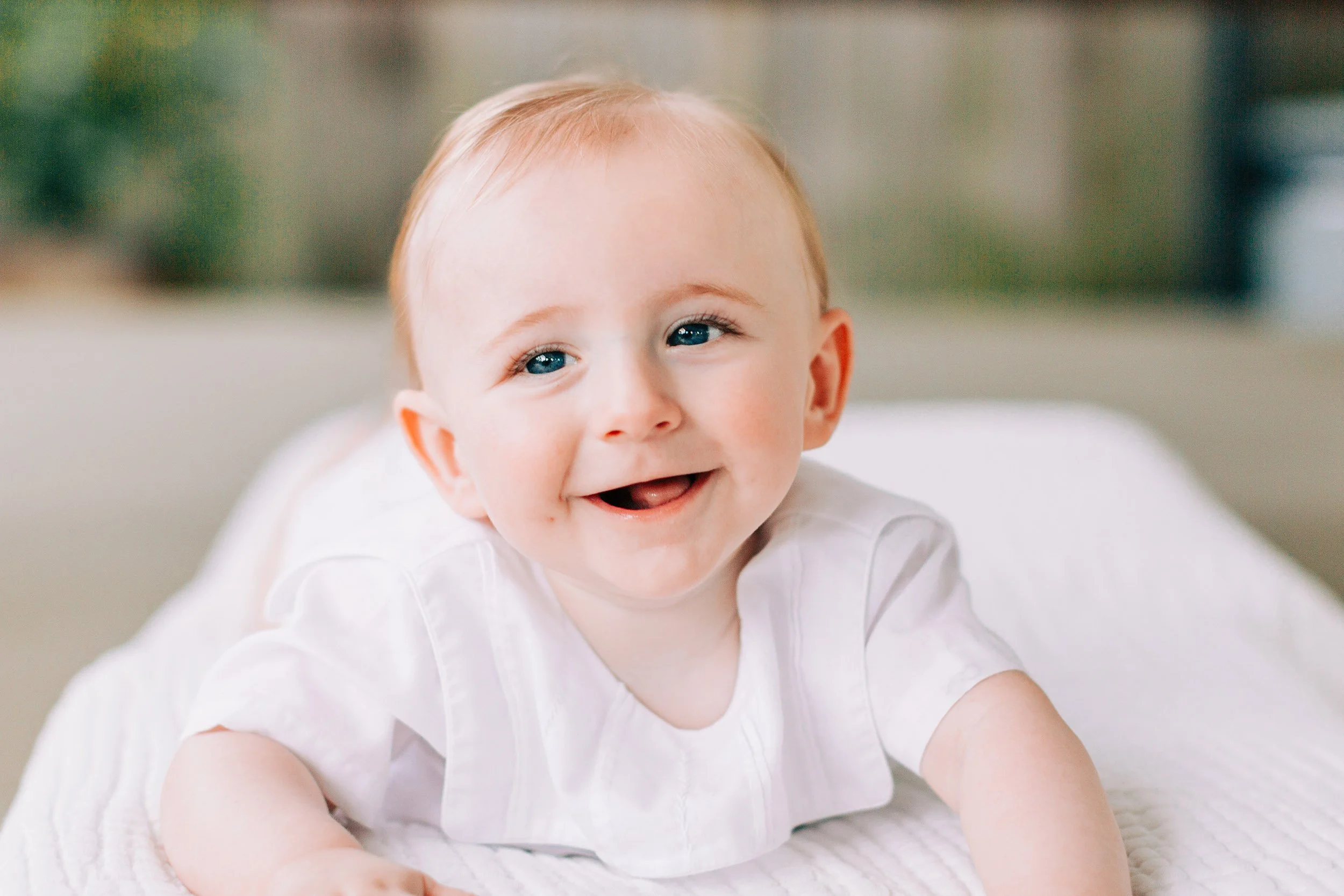 A smiling baby with blue eyes and light hair lying on a soft white surface, looking happy and playful.