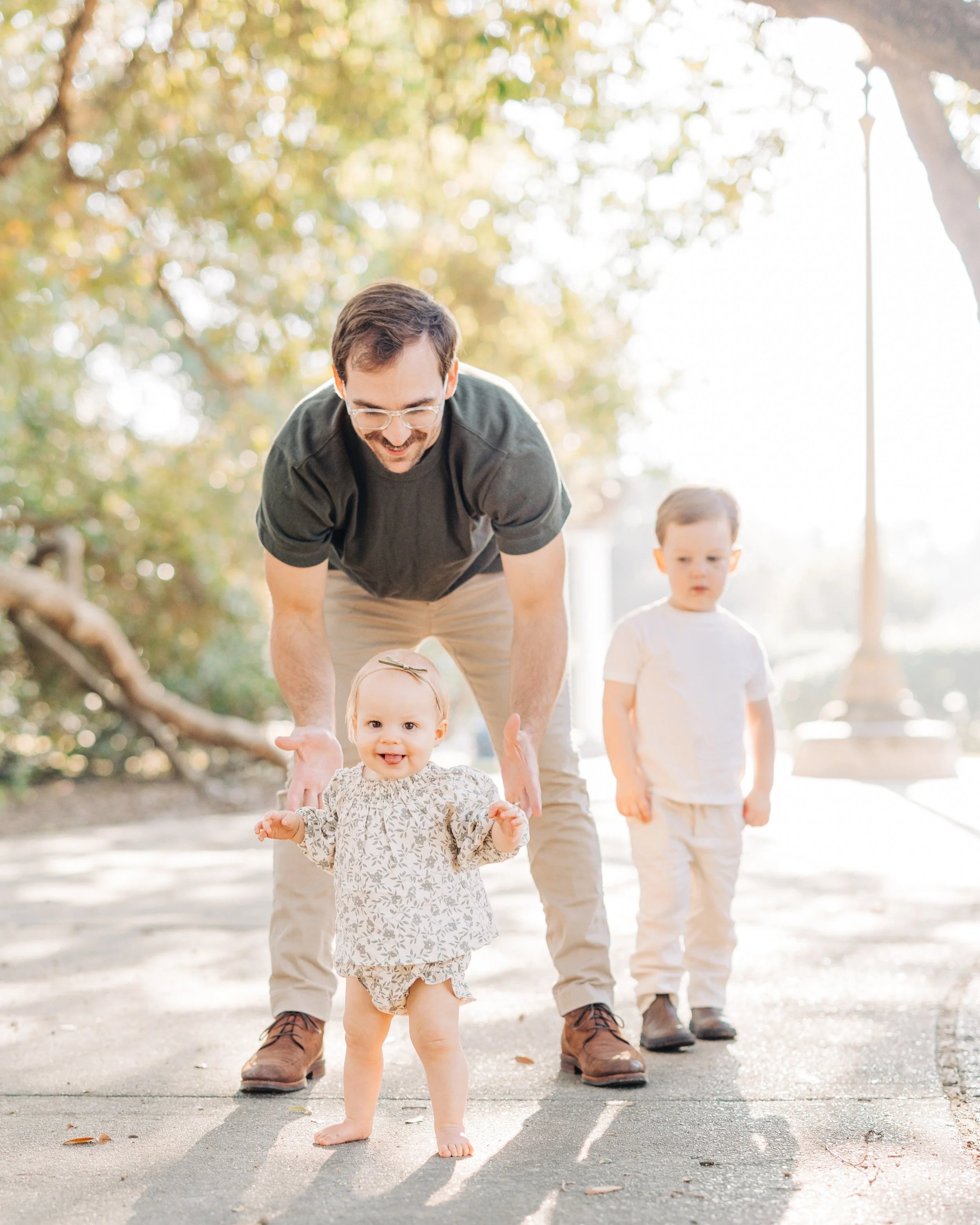 Father helping baby take first steps during candid family session outdoors in Baton Rouge