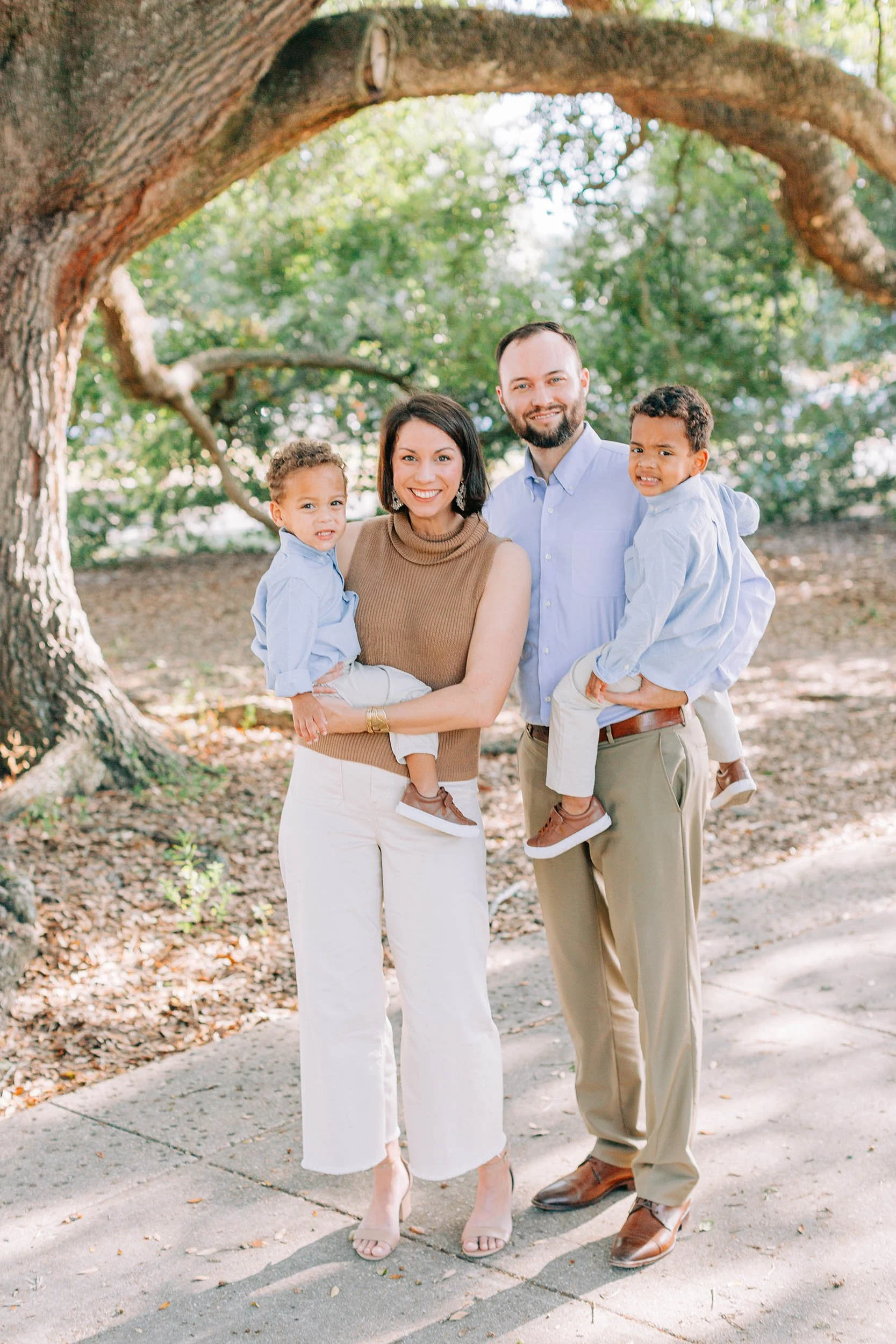 A diverse family of five standing outdoors under a large tree, smiling, with two young boys and a woman, all dressed in light-colored clothing.