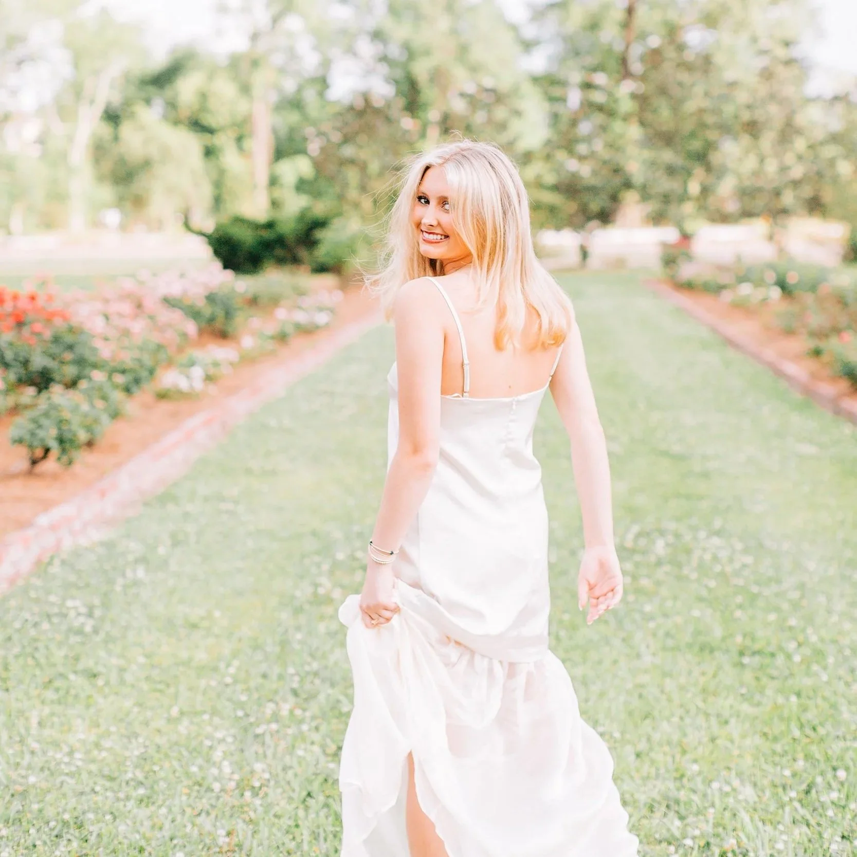 A young woman with blonde hair and a white dress walking outdoors in a park with trees and flowers, smiling and looking back over her shoulder.