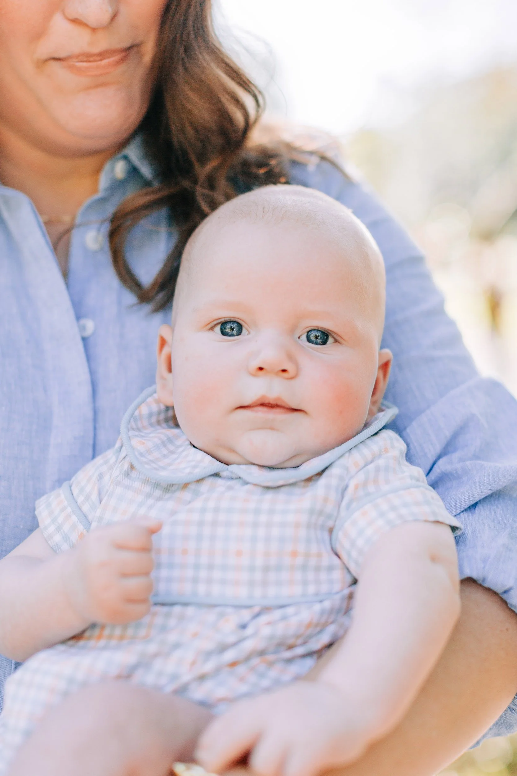 Close-up of a baby with blue eyes and a woman holding the baby, outdoors with blurred background. The woman is wearing a blue shirt, and the baby is dressed in a checked outfit.