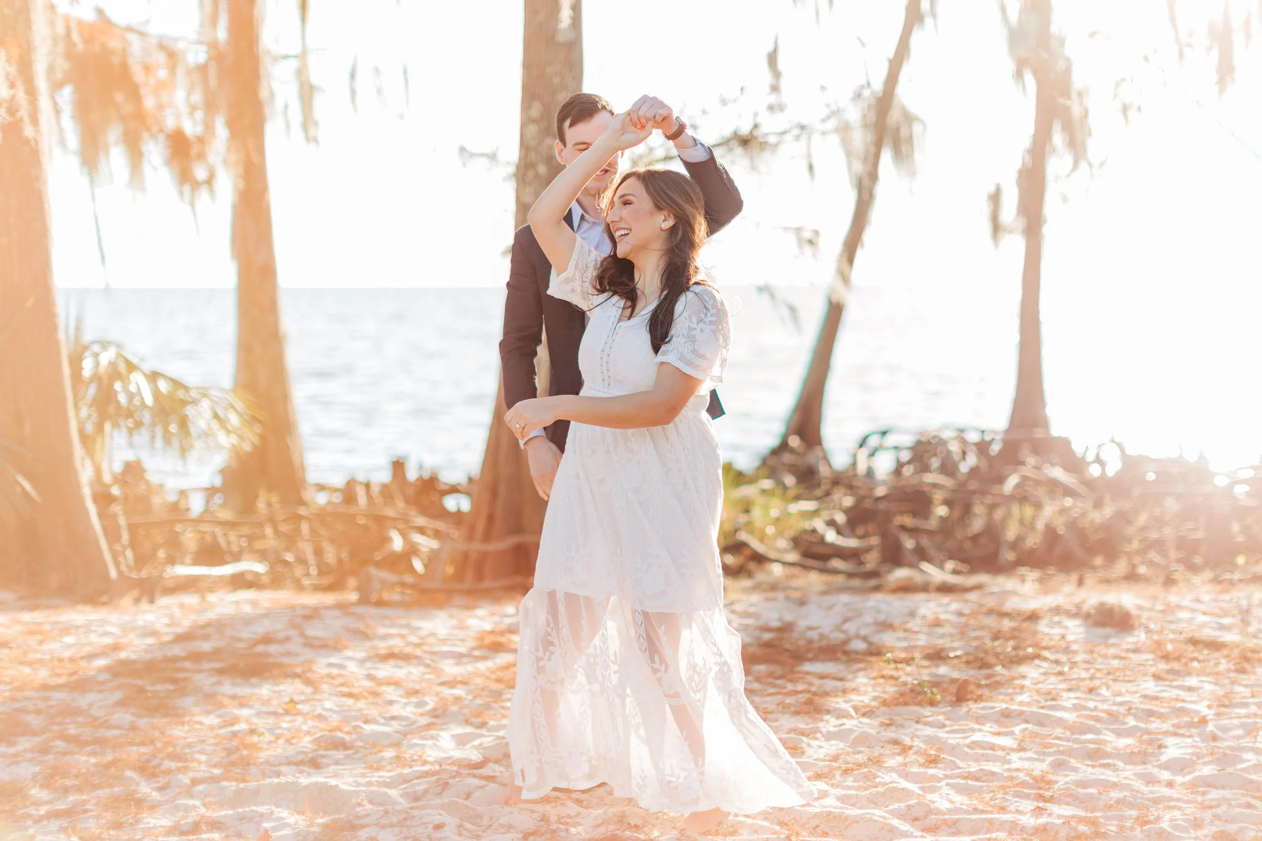 A couple dancing outdoors on a sunny day near the water, with the woman in a white lace dress and the man in a dark suit, surrounded by trees.