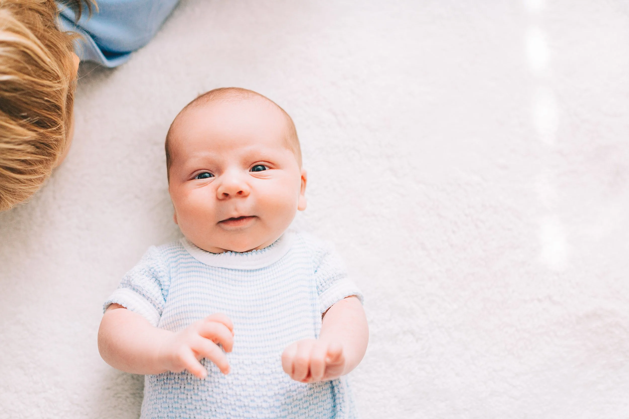 A baby boy with blue eyes and light skin lying on a soft white surface, wearing a white and blue striped onesie, looking up with a slight smile, with an adult partially visible beside him.