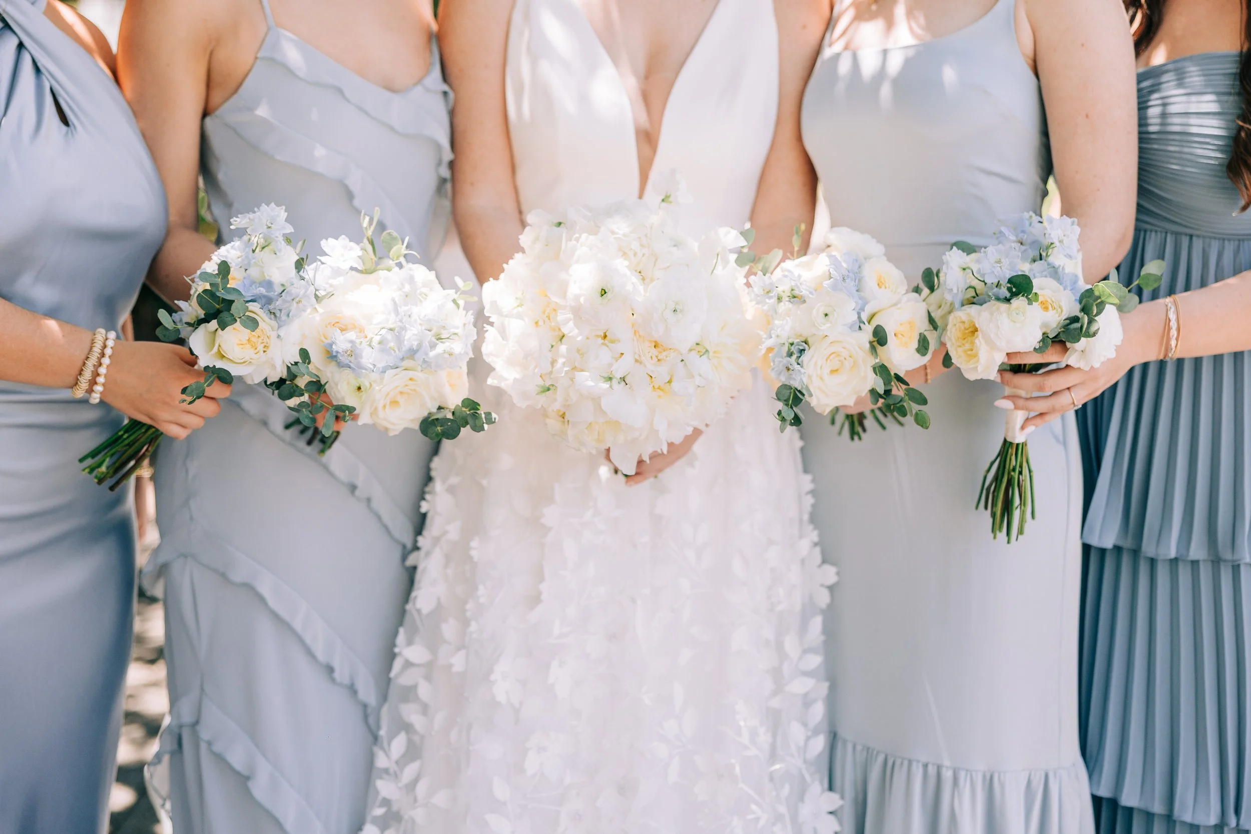 A bride in a white wedding dress holding a bouquet of white flowers and four bridesmaids in pastel-colored dresses, each holding a similar bouquet.