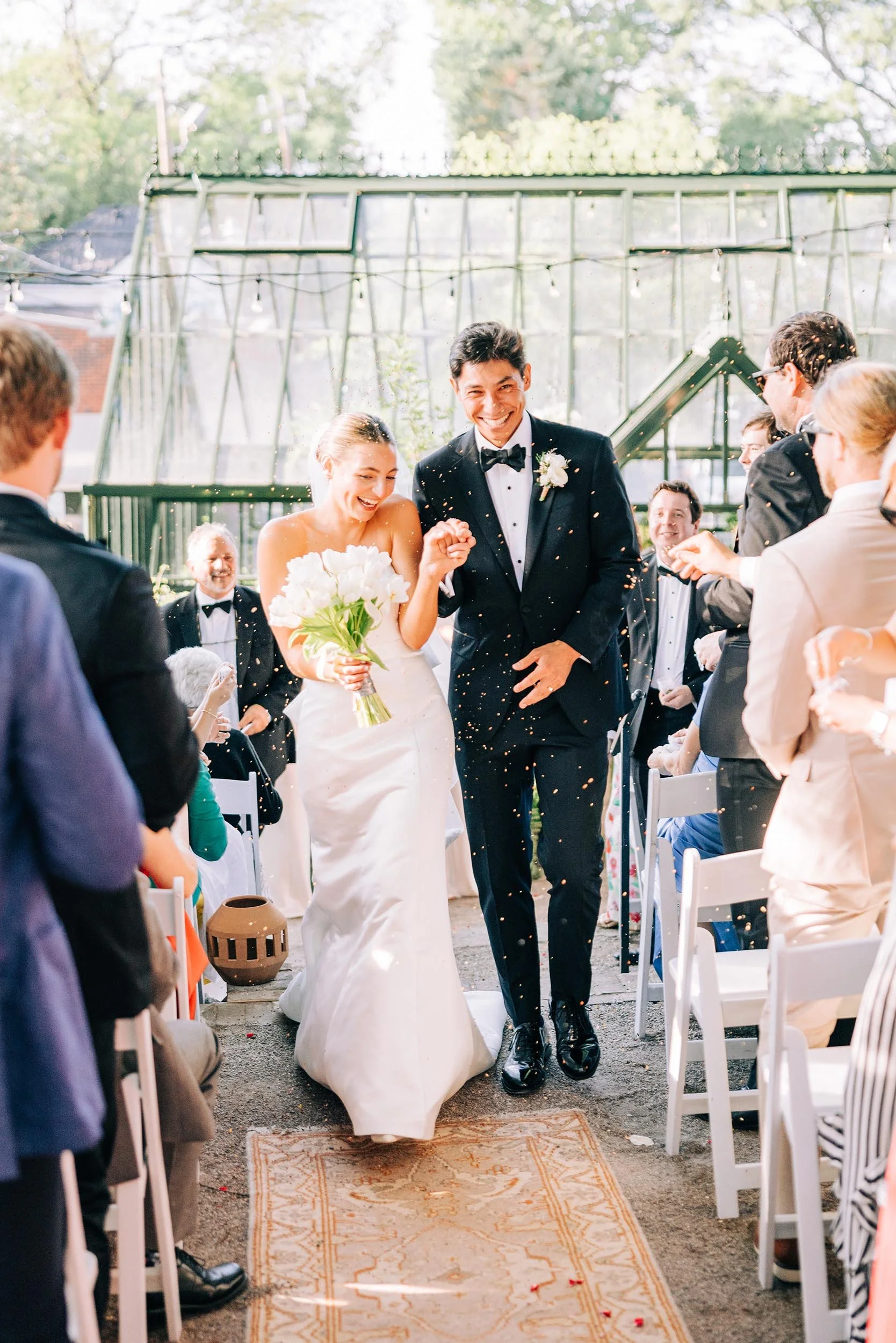A bride and groom smiling as they walk down an aisle lined with white chairs at an outdoor wedding ceremony, with guests on either side and string lights overhead.