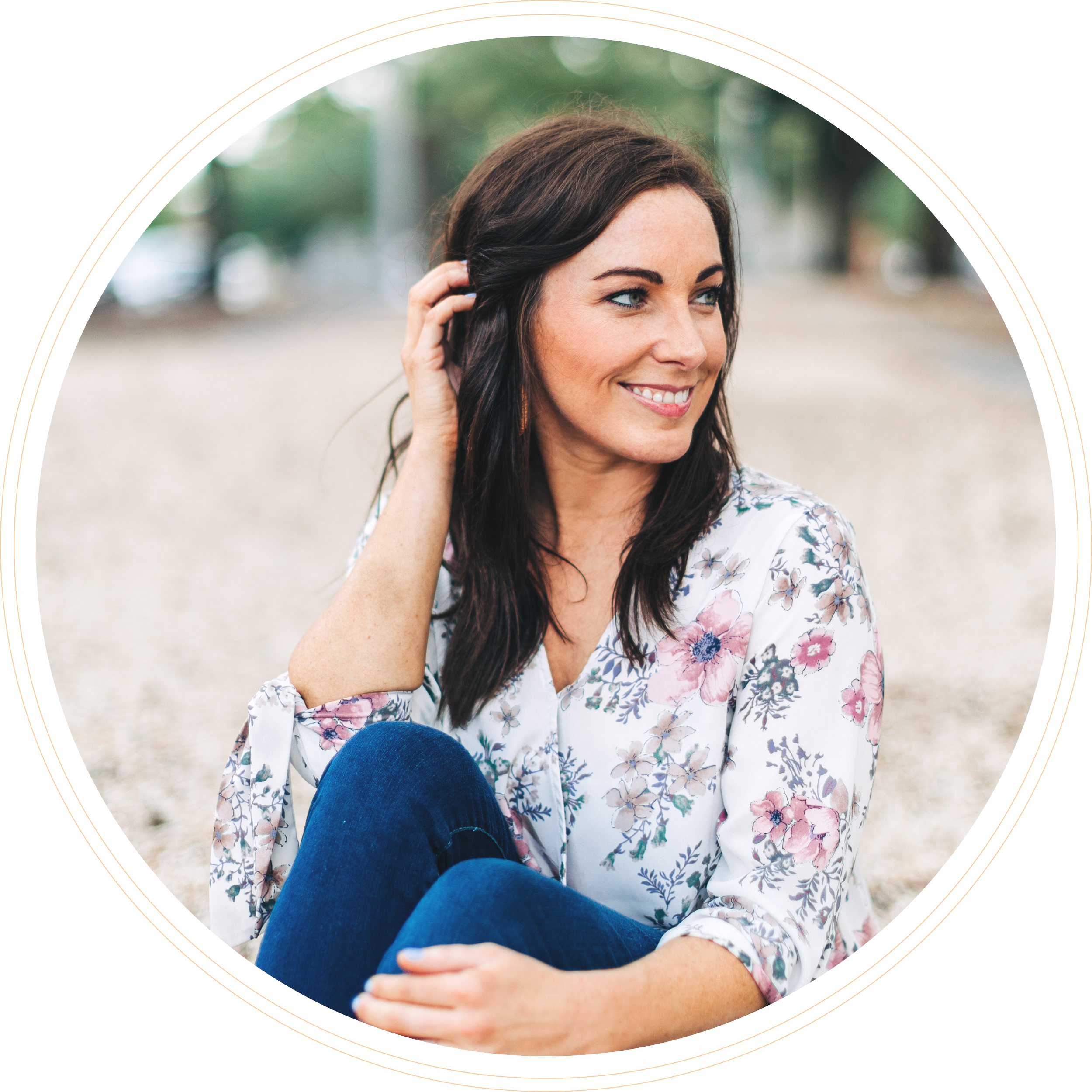 A woman with dark brown hair and blue eyes smiling outdoors, wearing a floral blouse, with her hand touching her hair. A person in blue jeans is seated with crossed legs in front of her.