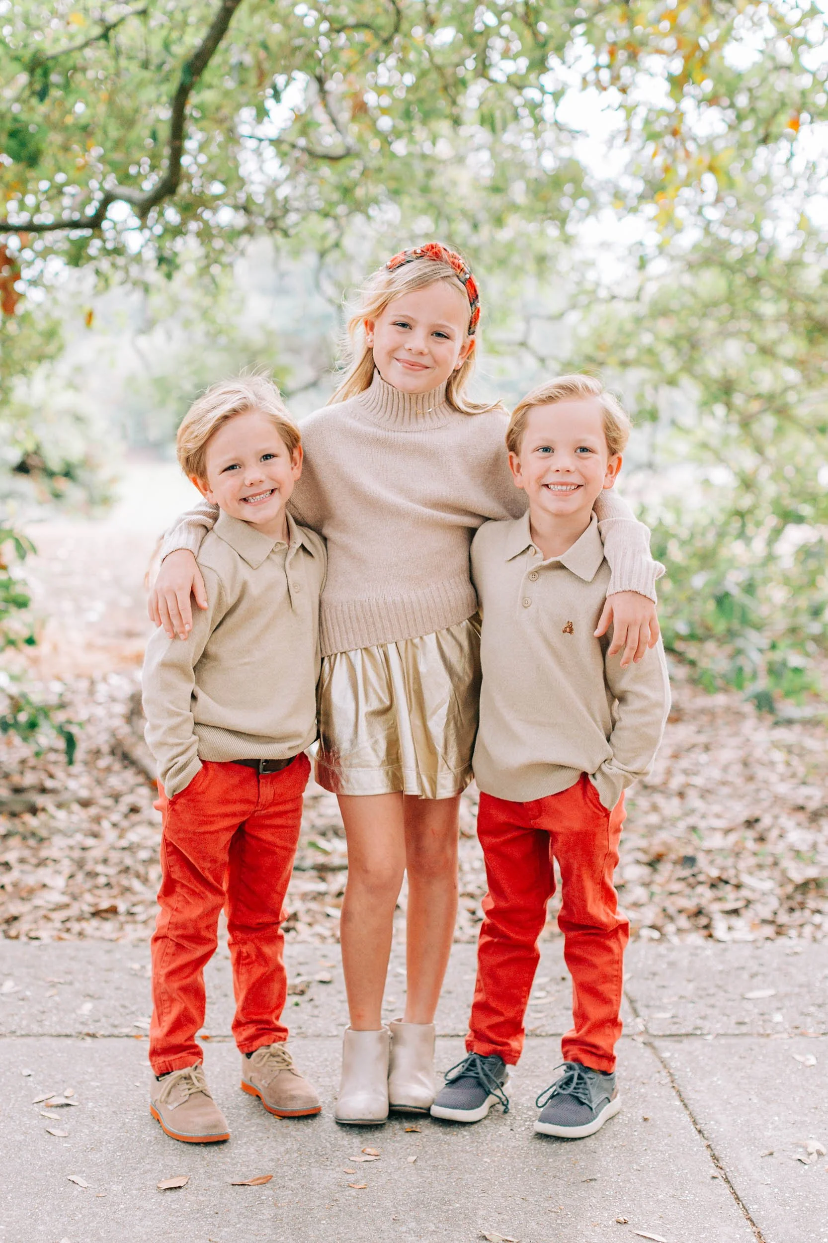 A girl and two boys standing close together outdoors, smiling. The girl has blonde hair, a red headband, a beige turtleneck sweater, and a golden skirt. The boys have blonde hair, beige polo shirts, and bright red pants. They are on a sidewalk with f