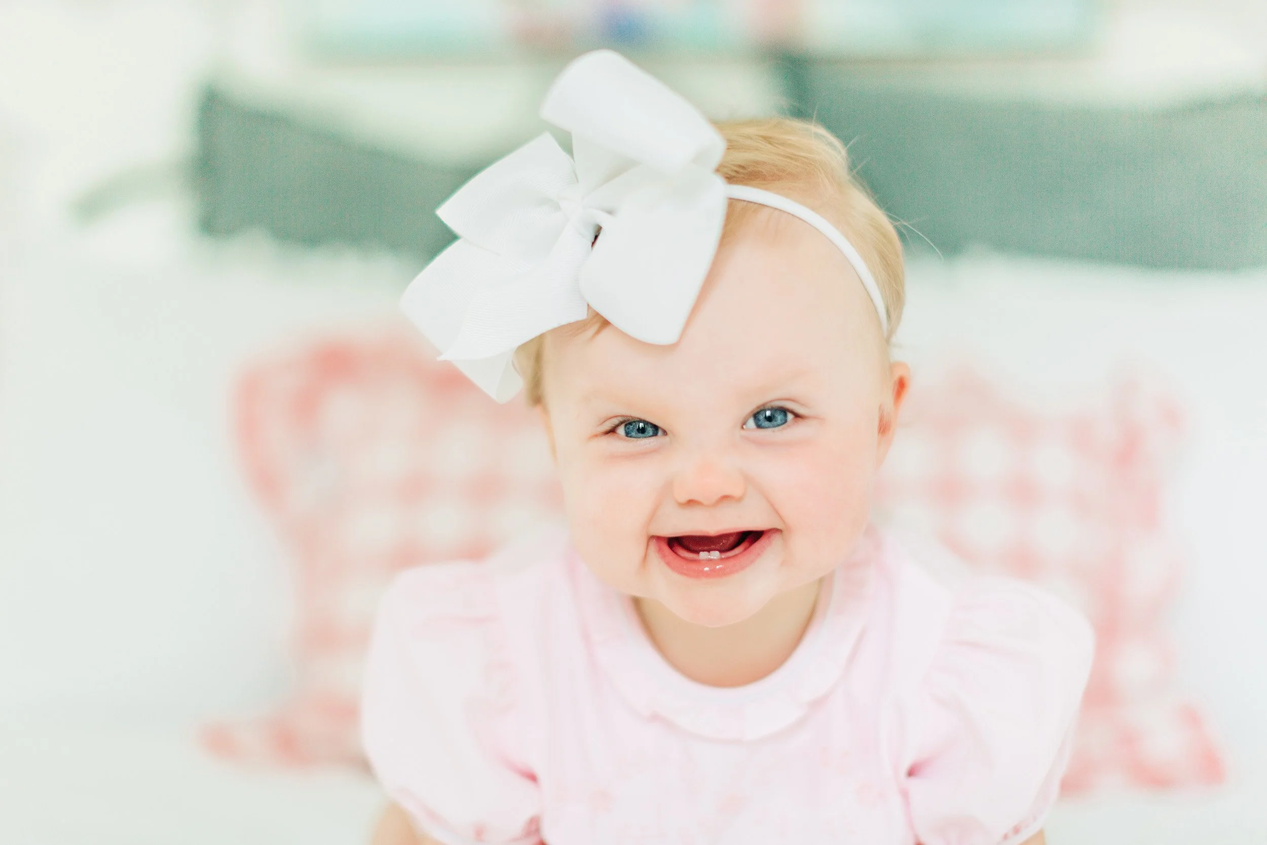 A smiling young girl with blue eyes and blonde hair, wearing a white headband with a large bow, reaching towards the camera.