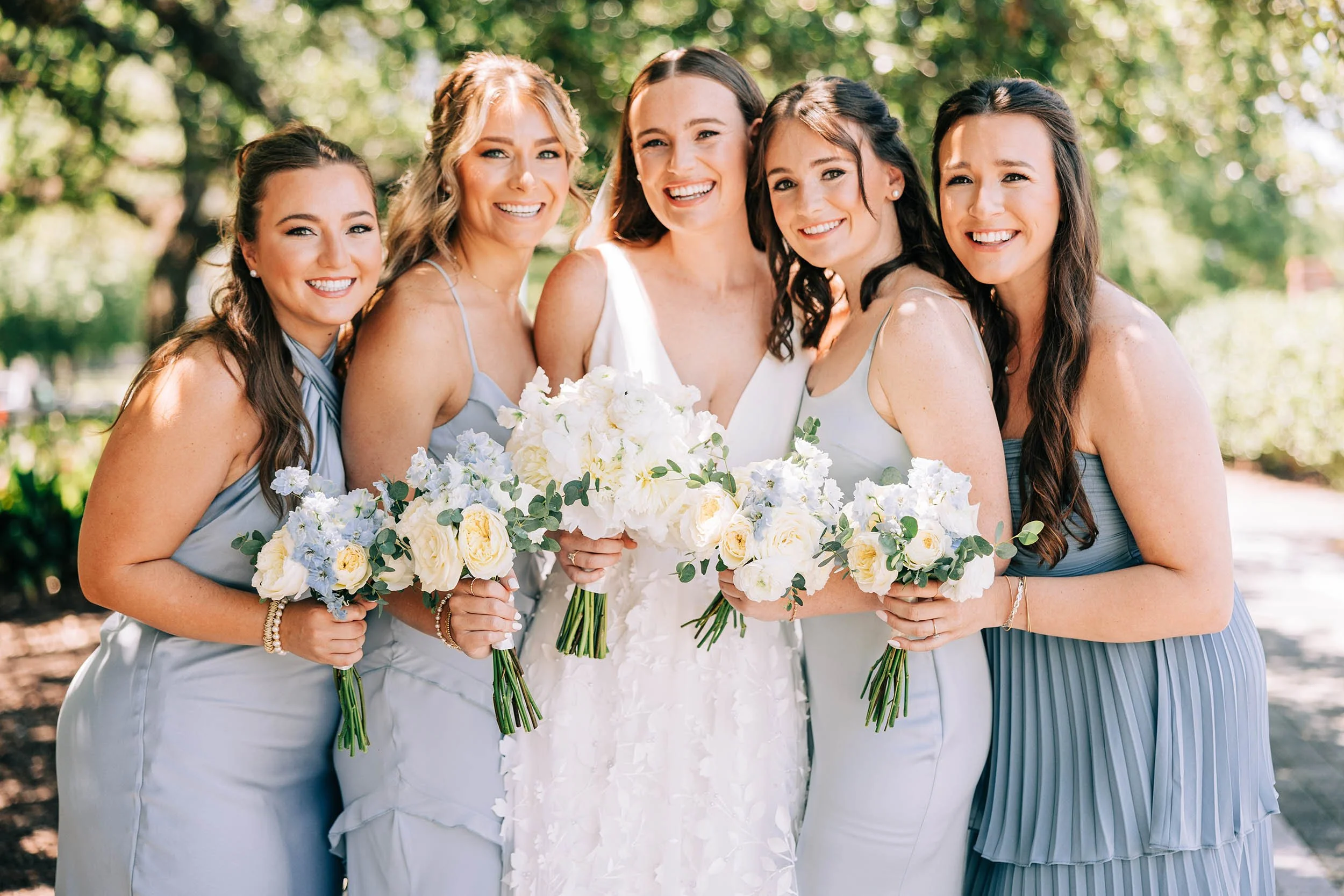 A group of six women standing outdoors, dressed in light-colored dresses, smiling and holding bouquets of white and light blue flowers, in a park with green trees.