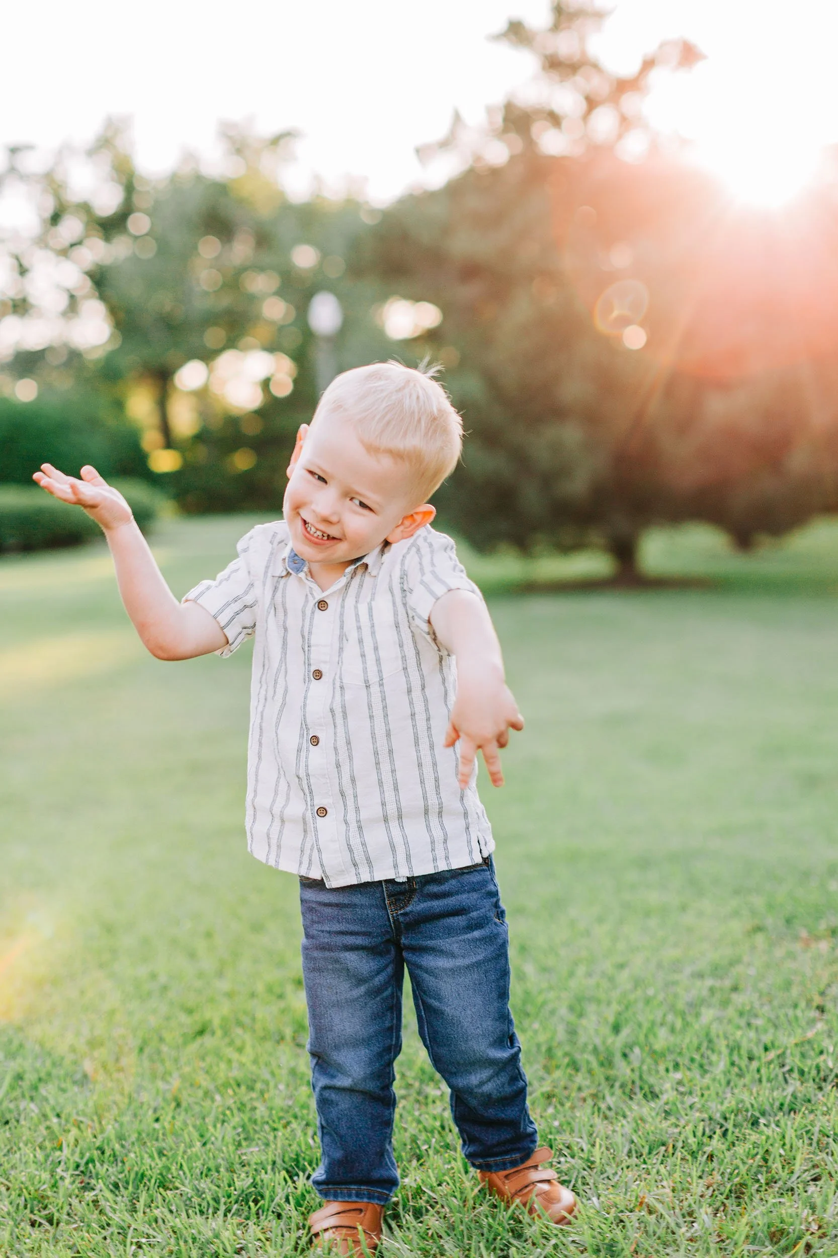 A smiling young boy with blonde hair stands outdoors on grass, wearing a striped shirt, blue jeans, and brown shoes, with trees and sunlight in the background.