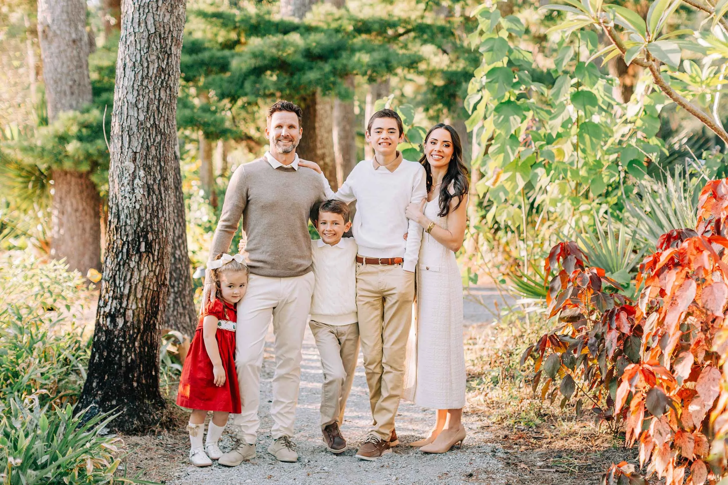 A family of six smiling and posing together outdoors in a park-like setting with trees and colorful foliage.