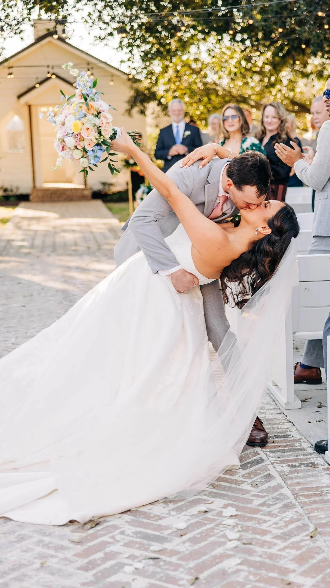 A bride and groom sharing a kiss during their outdoor wedding ceremony. The groom is leaning over the seated bride, who is holding a colorful bouquet of flowers, in front of a small church-like building. Guests stand in the background, clapping and smiling.