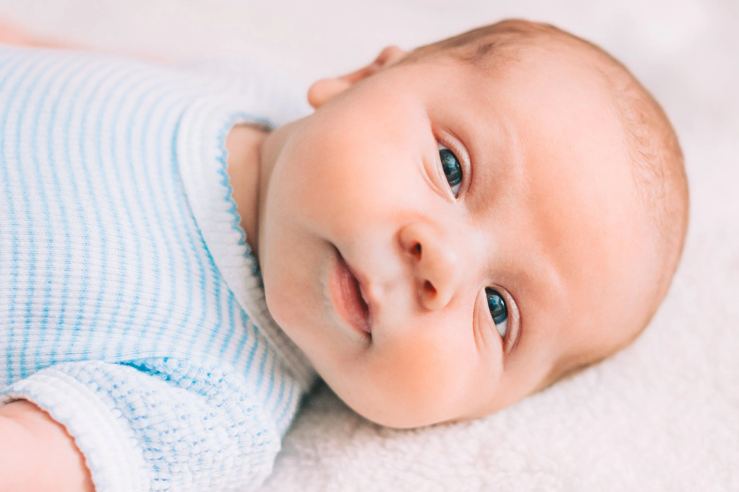 Close-up of a baby's face lying on a soft surface, wearing a light blue and white striped onesie, with blue eyes and light hair.