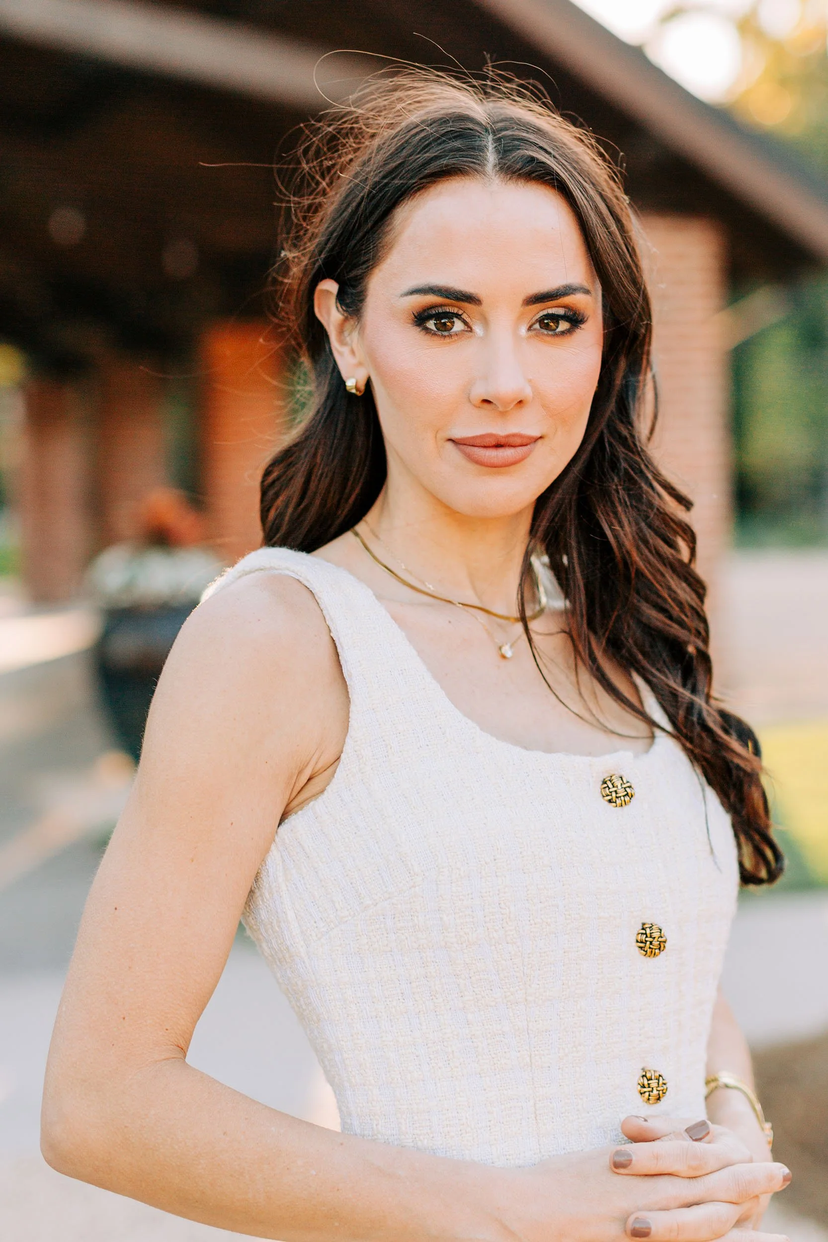 A woman with long, dark brown hair, wearing a white sleeveless dress with decorative buttons, standing outdoors near a building with a blurred background of trees.