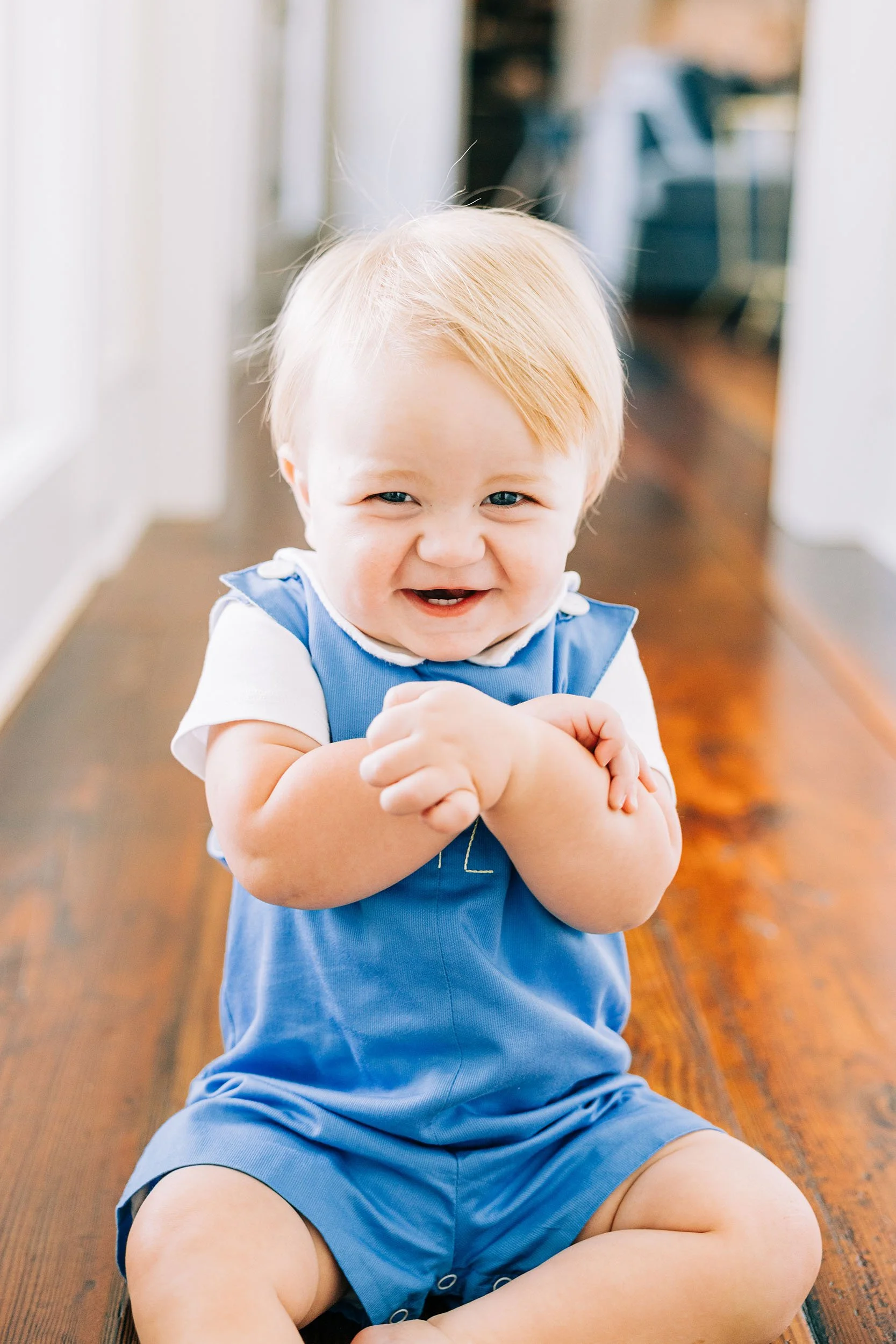 A young child with blonde hair and blue eyes, sitting on a wooden floor, smiling, wearing a blue overall and white shirt.