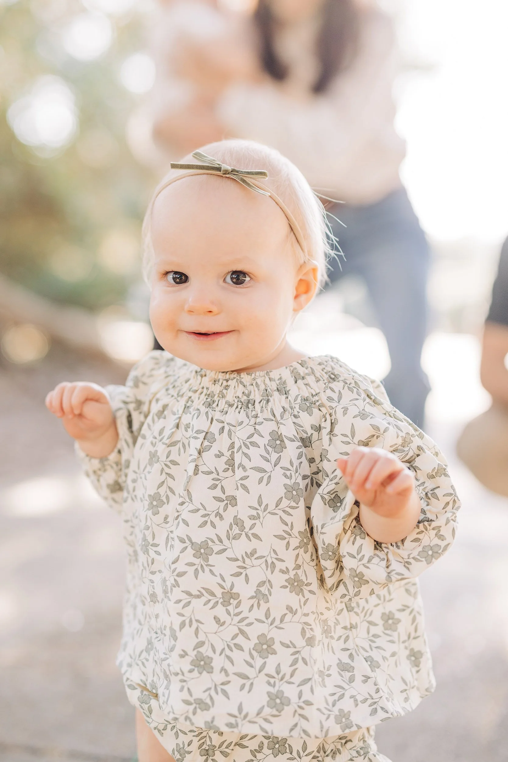 A young girl with blonde hair wearing a beige floral dress and a beige headband with a bow, smiling outdoors with blurred trees and sunlight in the background.