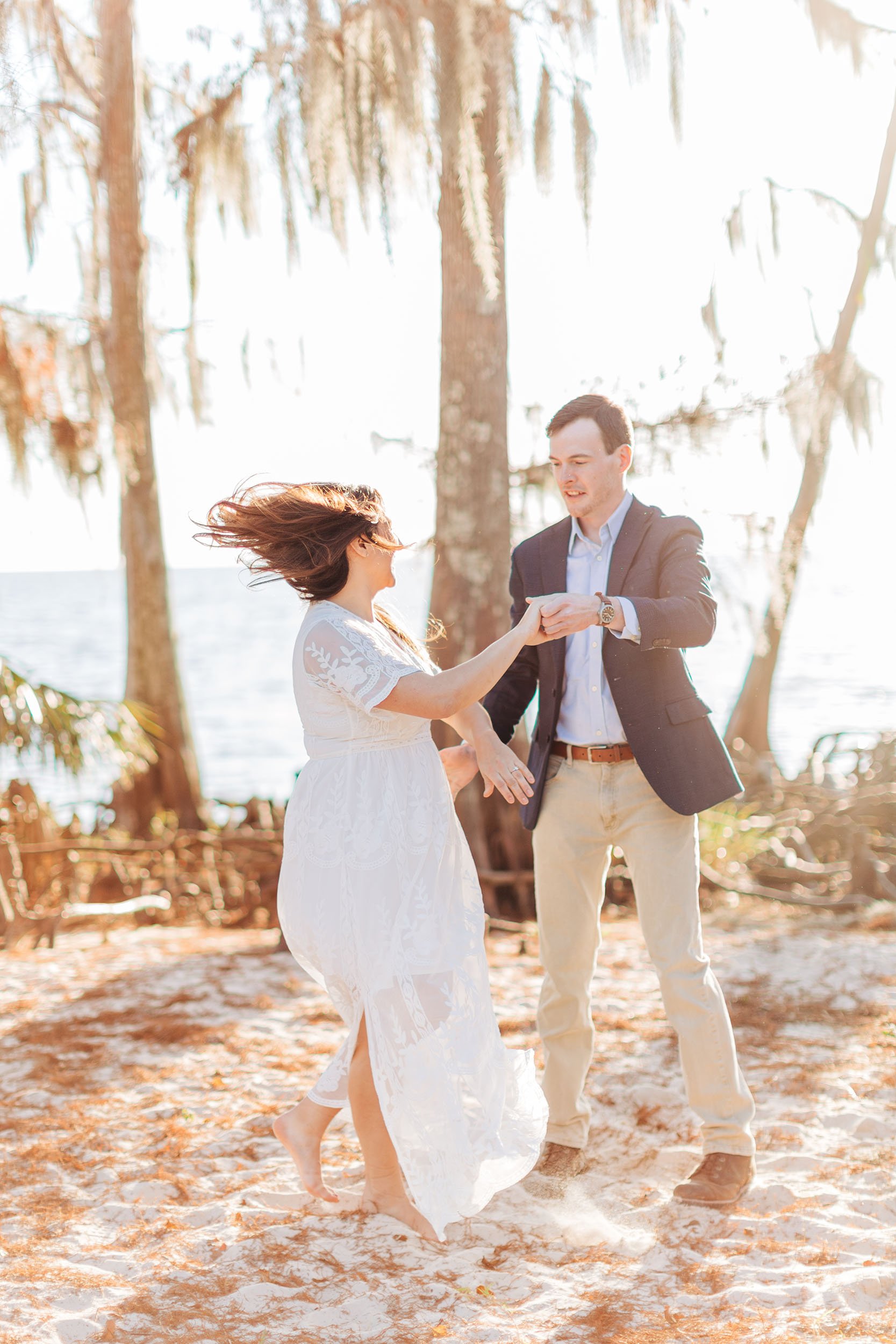 A couple getting married on a beach with trees and water in the background. They are holding hands and dancing, with the woman in a white lace dress and the man in a dark blazer and light-colored pants. The wind is blowing the woman's hair.
