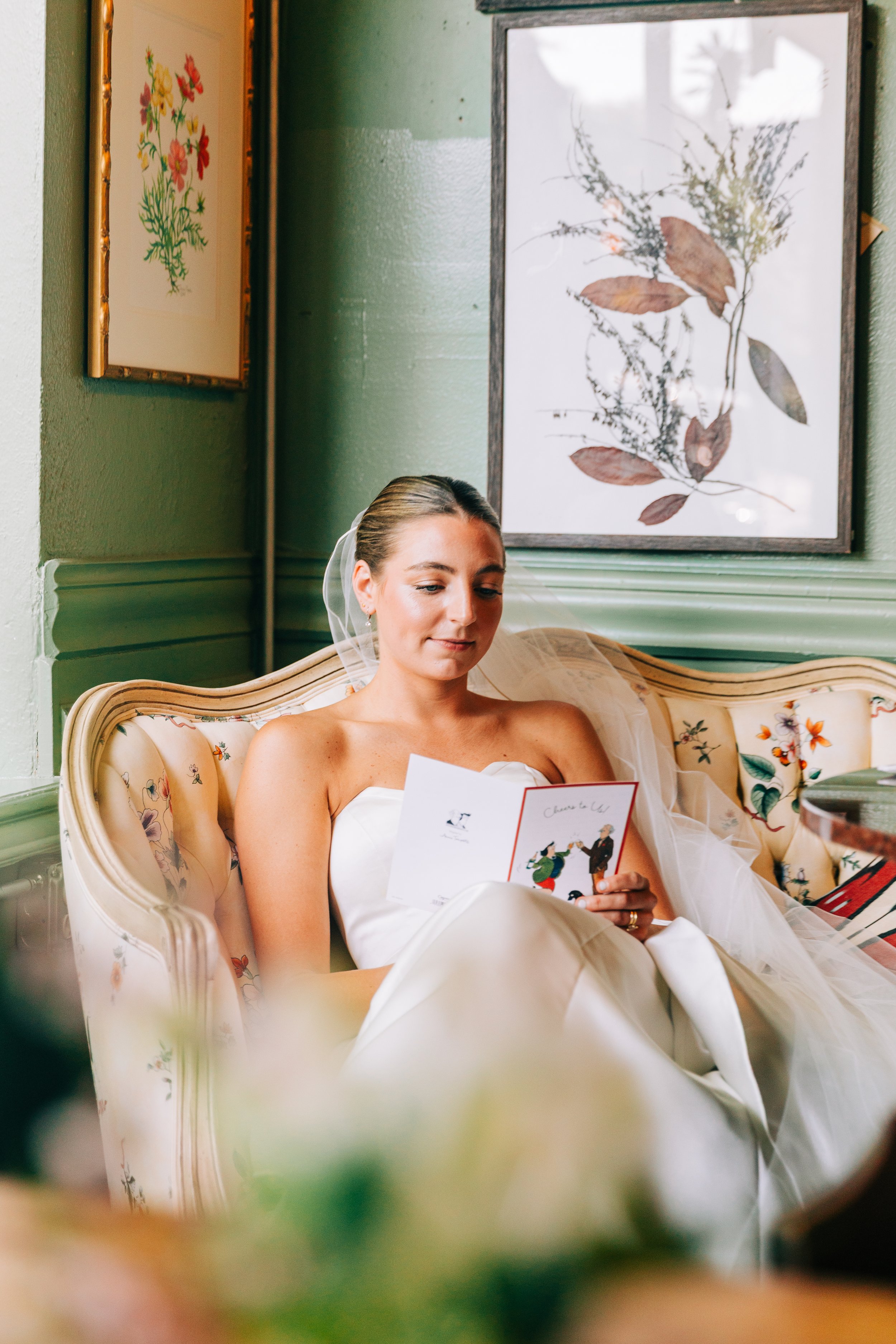 A bride with a veil and wedding dress, sitting on a floral-patterned vintage sofa, reading a card, with framed botanical art on a green wall behind her.