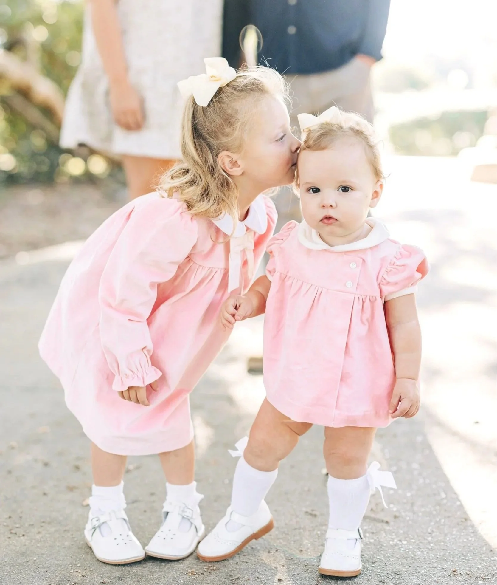Close-up of two young girls dressed in pink outfits, wearing white socks and sneakers, standing outdoors on a gravel surface, holding hands, with a blurred person in the background.
