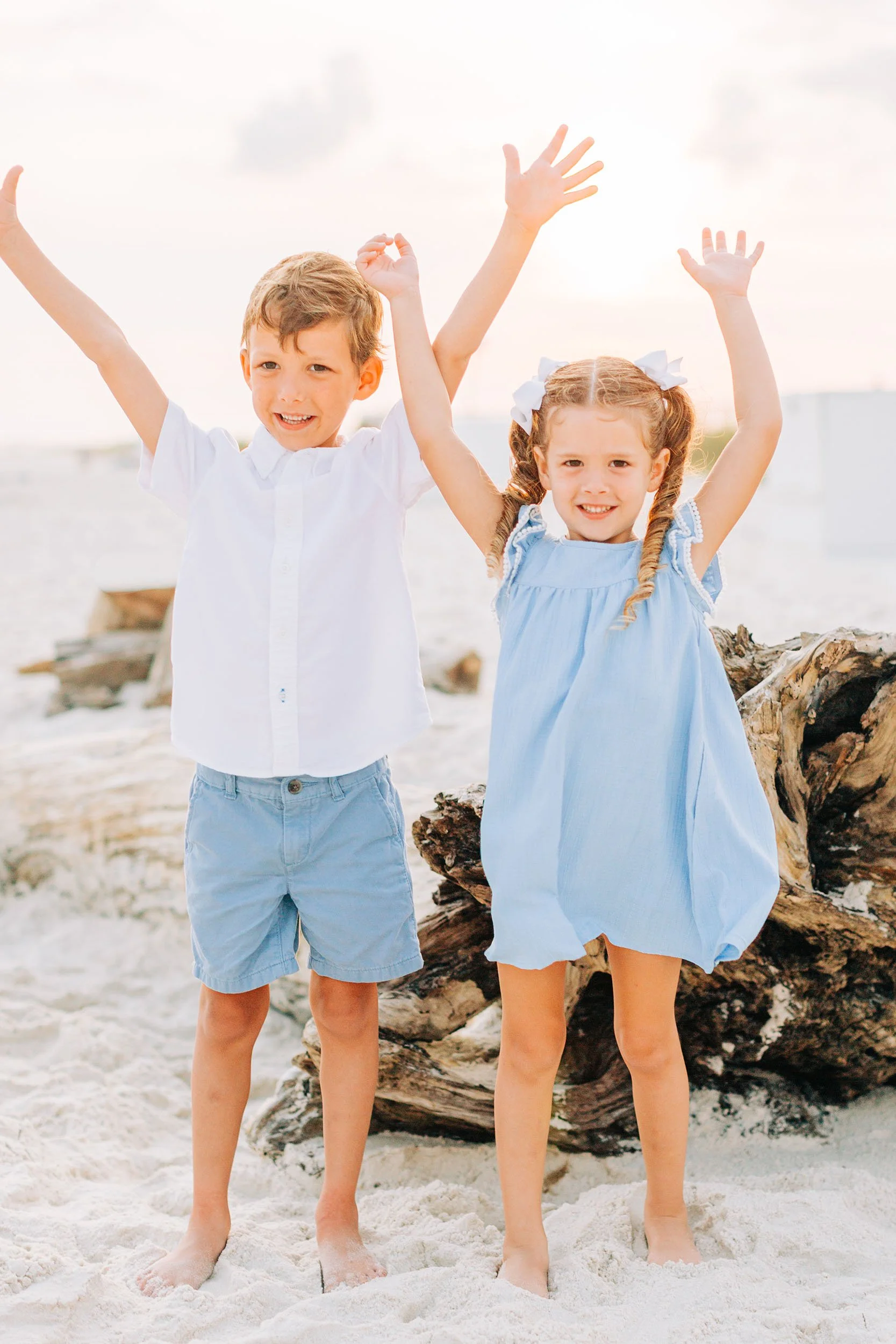 A boy and girl standing barefoot in the sand on a beach, raising their arms in the air, with a driftwood log and the ocean in the background.