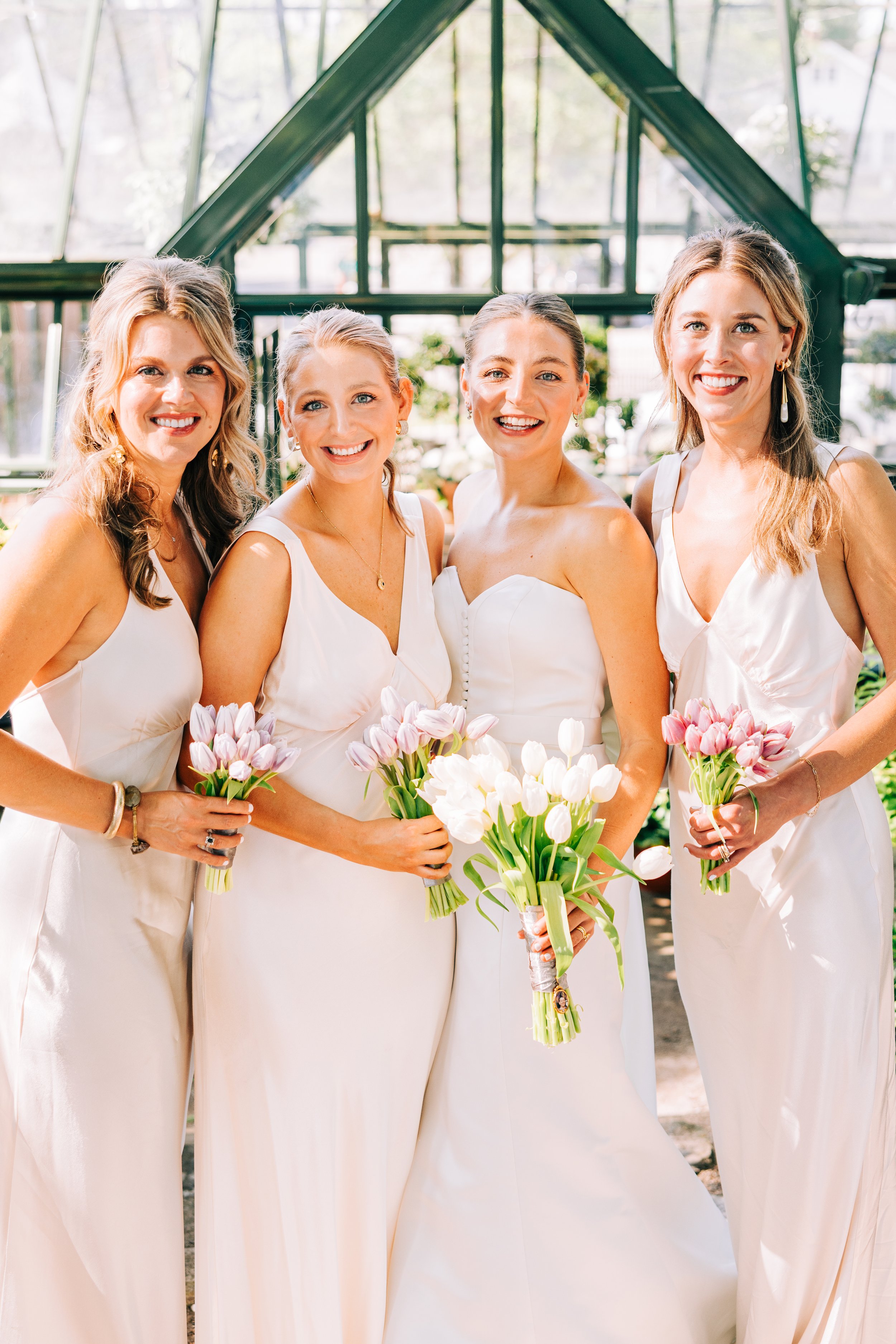 Group of four women wearing white dresses and holding pink and white tulip bouquets, standing together outdoors with a greenhouse structure in the background.