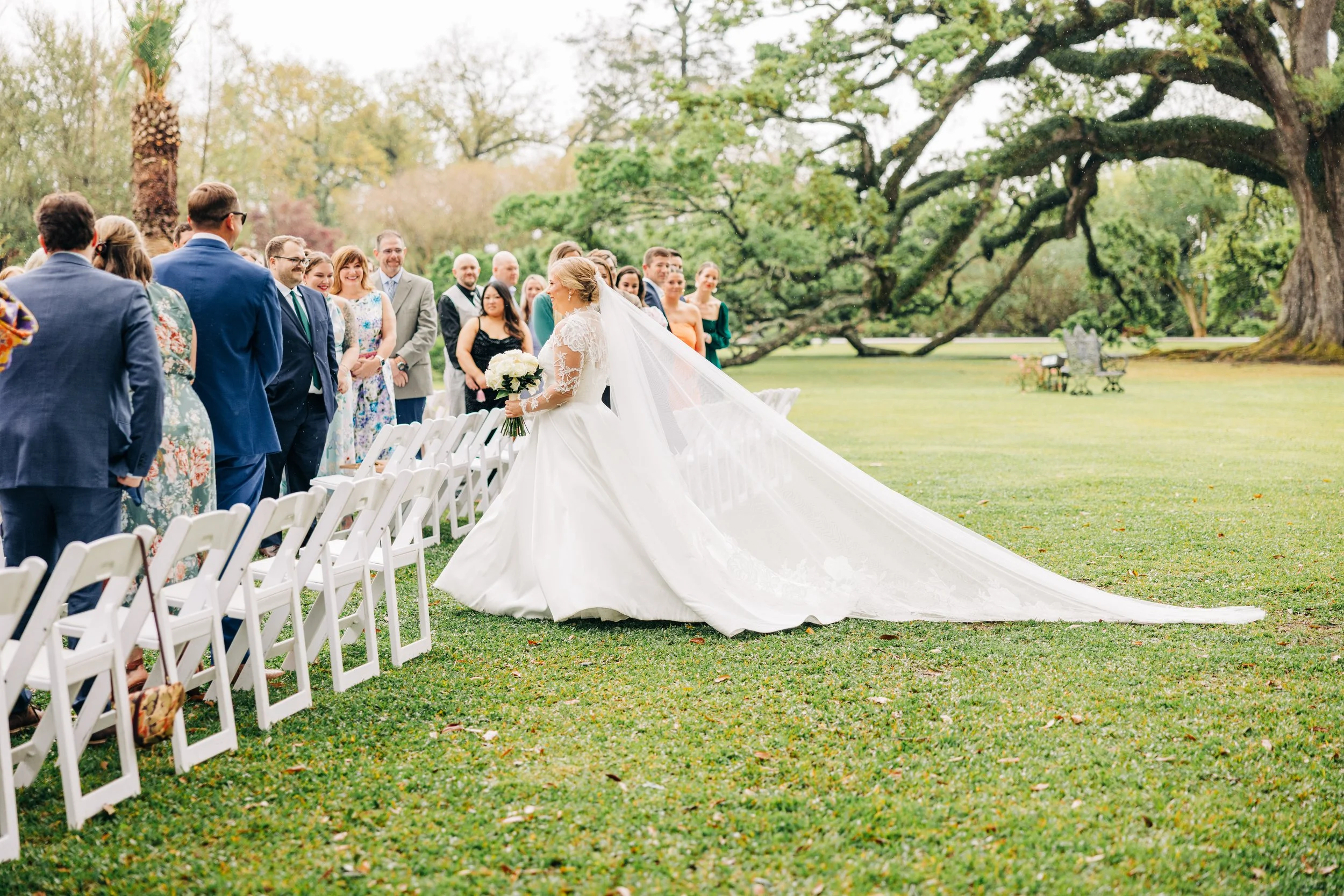 Bride in a white wedding gown holding a bouquet, standing and smiling at guests outside at a wedding ceremony with trees in the background.