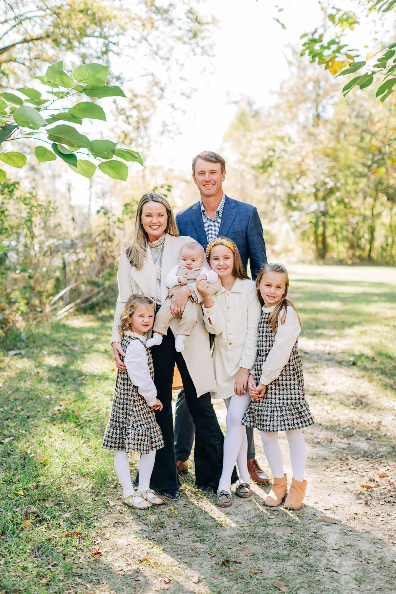 A family of seven, including two adults and five children, standing outdoors in a park on a sunny day with trees and greenery in the background.