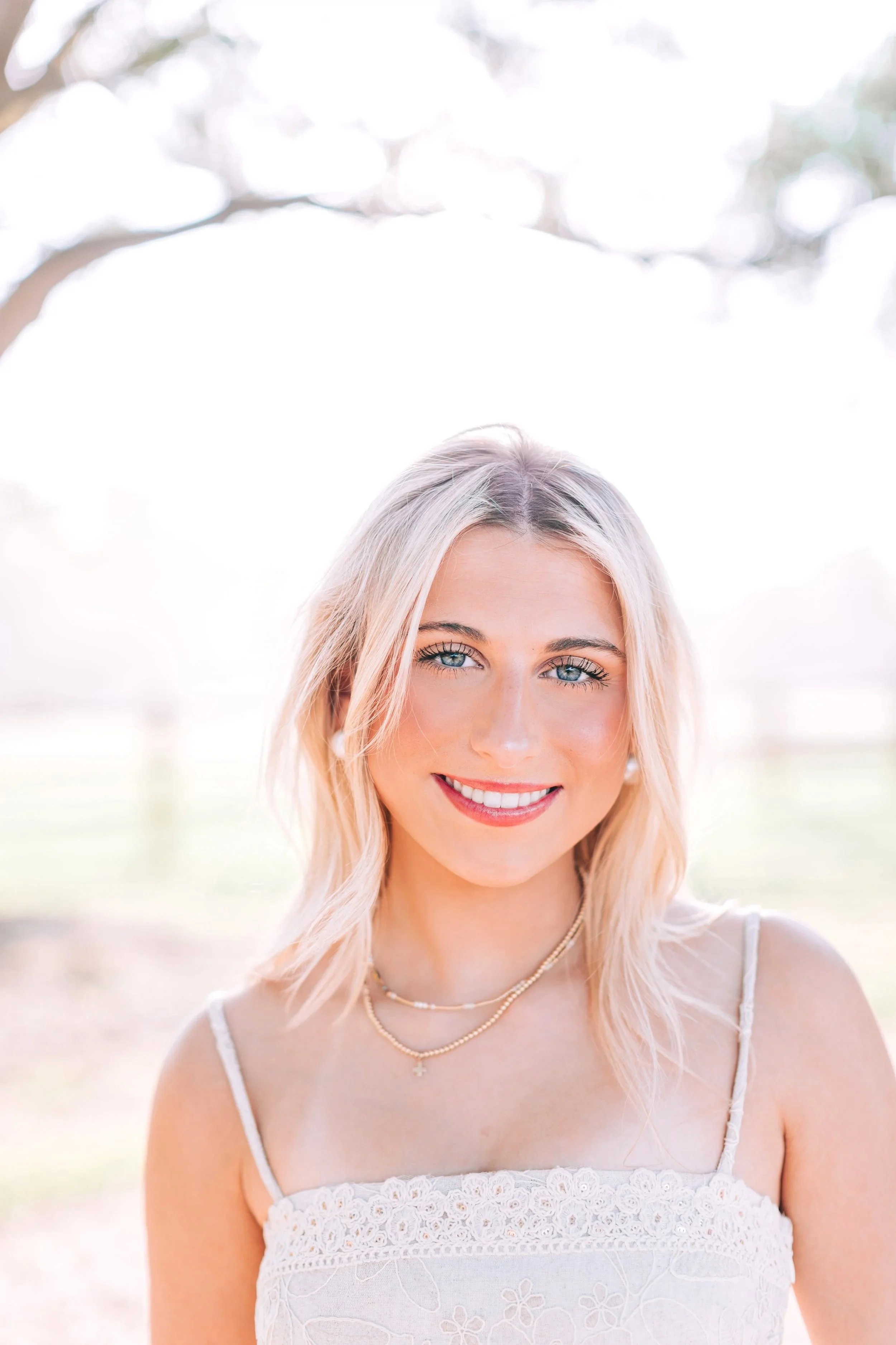 A woman with blonde hair and blue eyes smiling outdoors on a sunny day, wearing a white lace dress and gold necklaces.