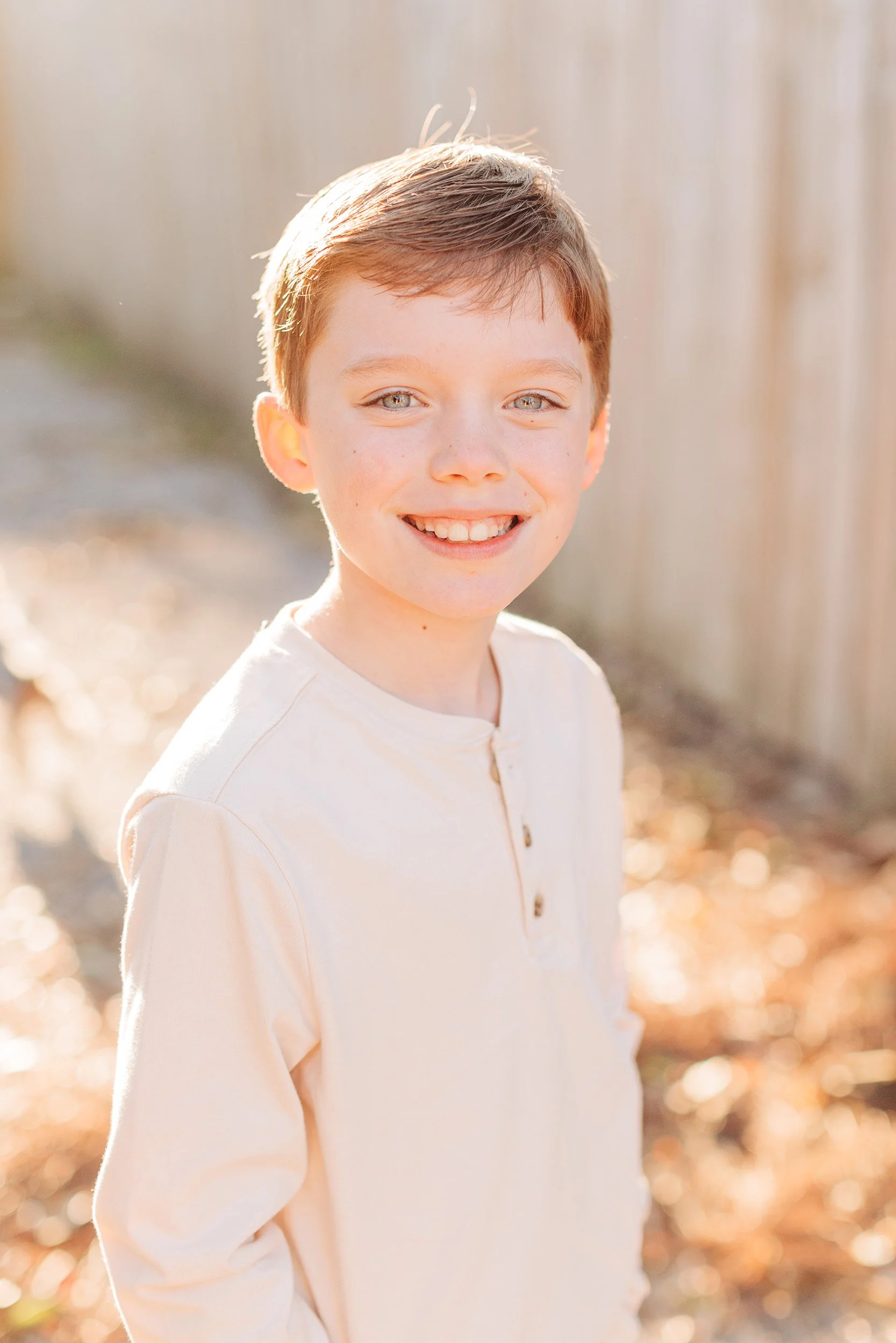 A young boy with brown hair and blue eyes smiling outdoors with sunlight backlighting his face, wearing a beige shirt with buttons.