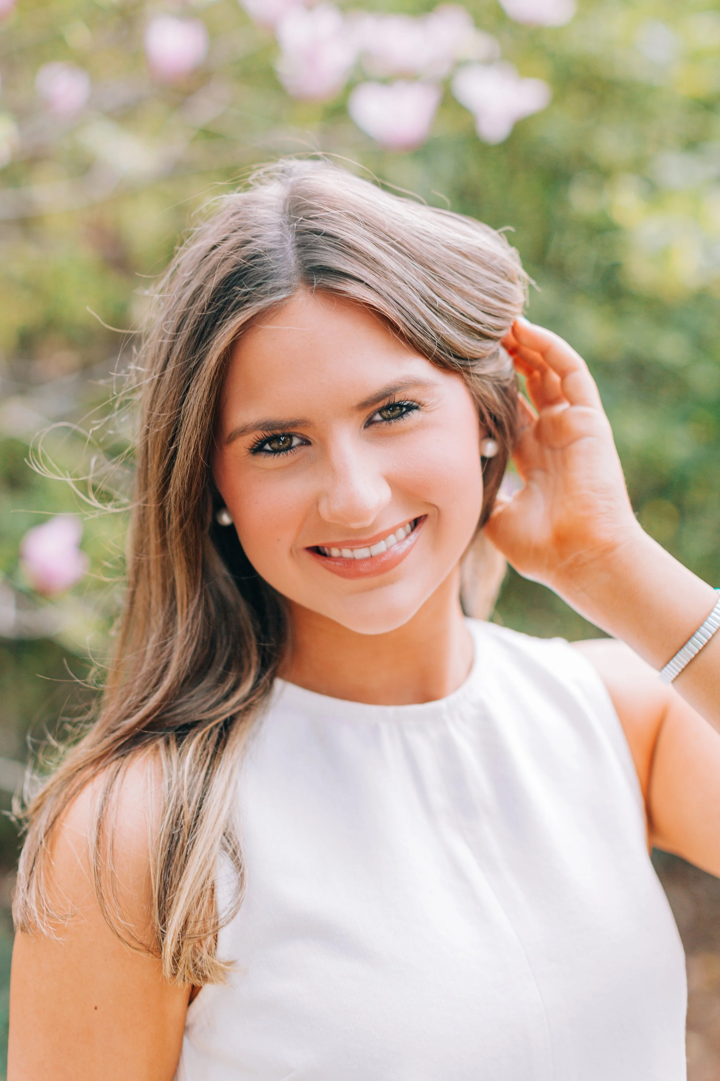 baton rouge senior portraits at burden gardens, brown haired girl smiling and brushing her hair behind her ears standing in front of a pink azalea bush, close up senior portrait