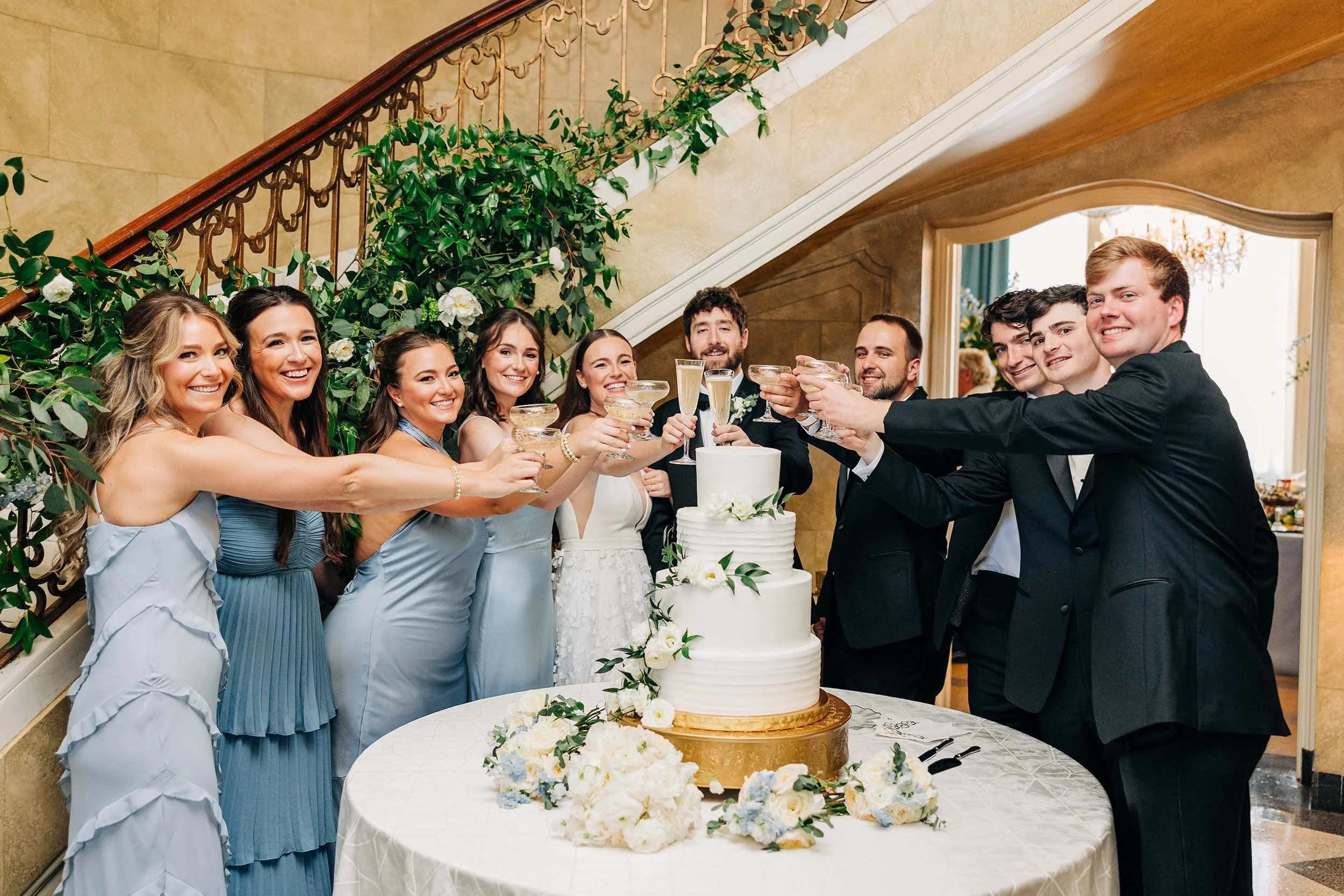A group of ten people, including women in light blue dresses and men in black suits, celebrating at a wedding reception with champagne glasses raised, a wedding cake, and floral decorations in a hallway with a staircase and chandelier.