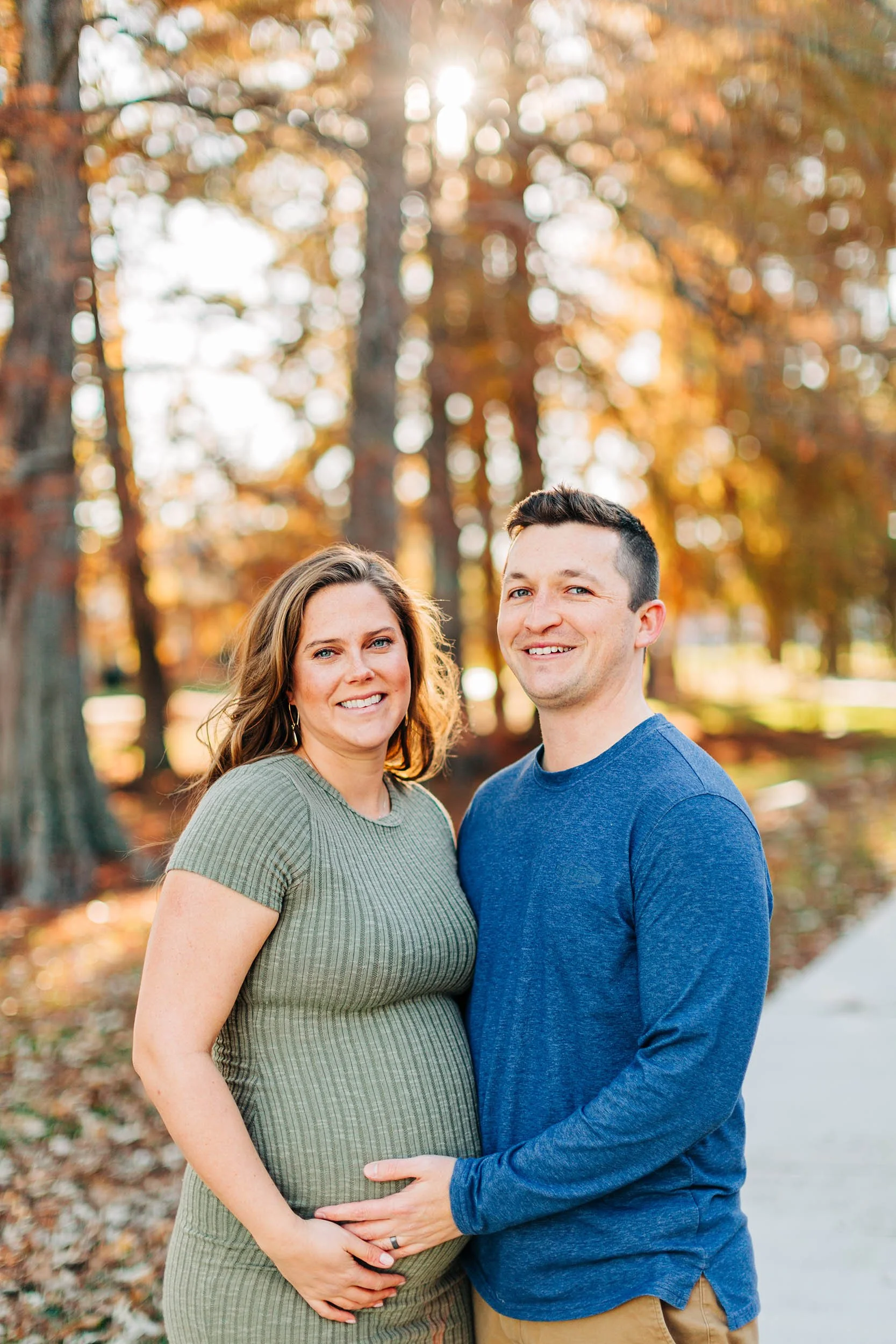 A pregnant woman and a man standing outdoors in a park with autumn leaves and trees in the background, smiling at the camera.