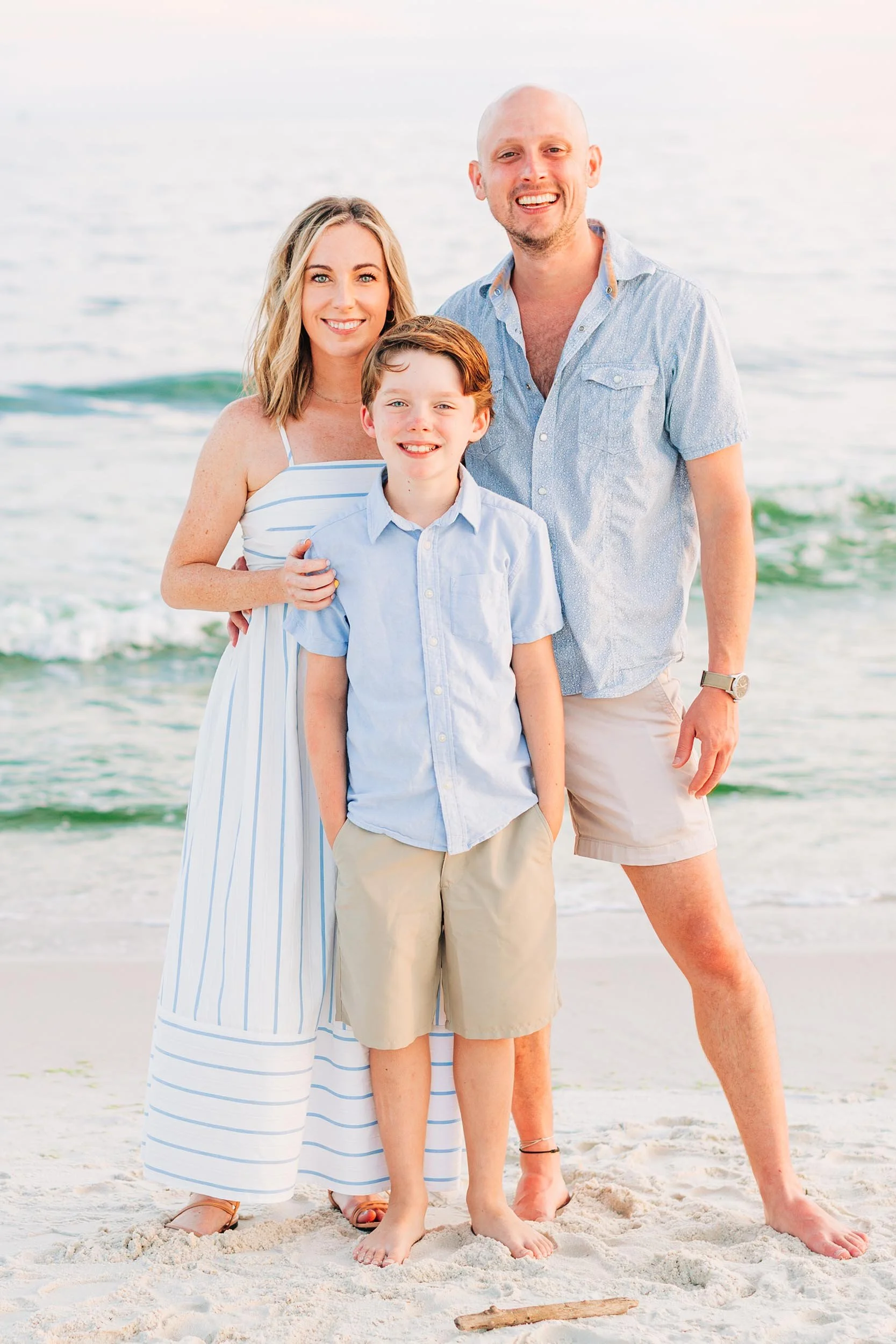 A family of three standing on the beach by the ocean, smiling. The woman has blonde hair and is wearing a white and blue striped dress. The man is bald, wearing a light blue shirt and beige shorts. The boy has brown hair, wearing a light blue shirt and khaki shorts.