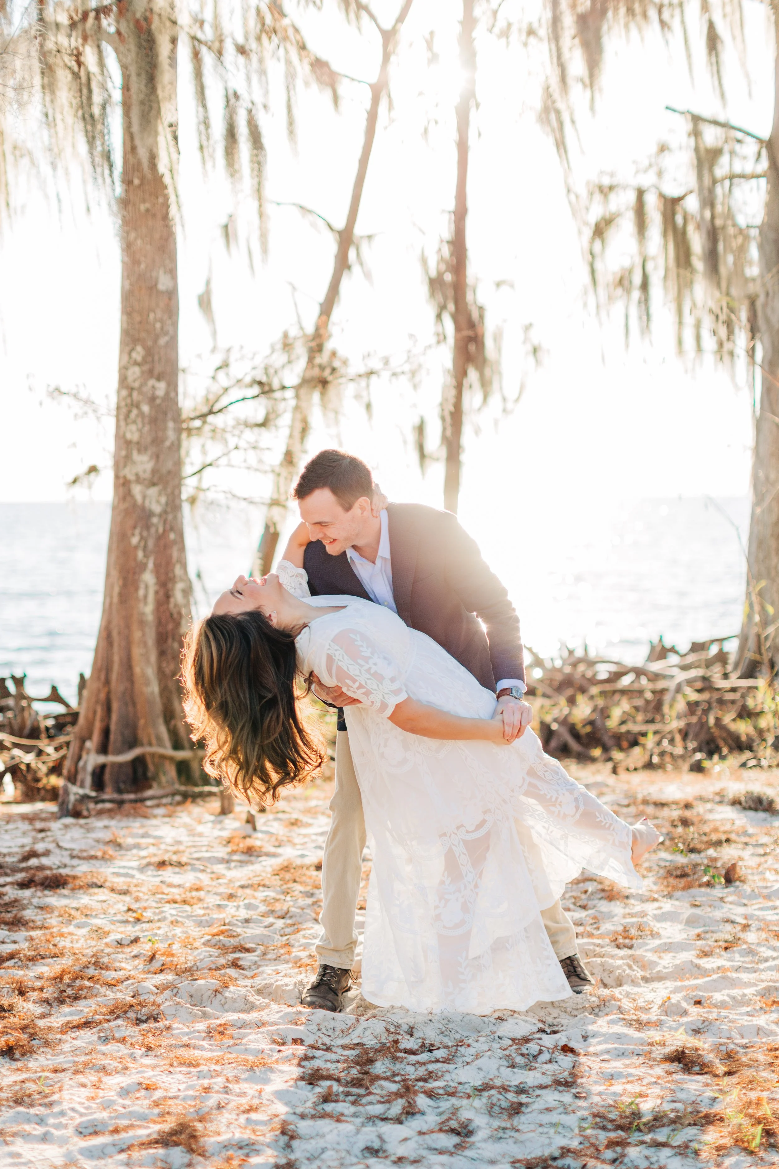 A couple dancing on a sandy beach with tall trees in the background, backlit by the setting sun.